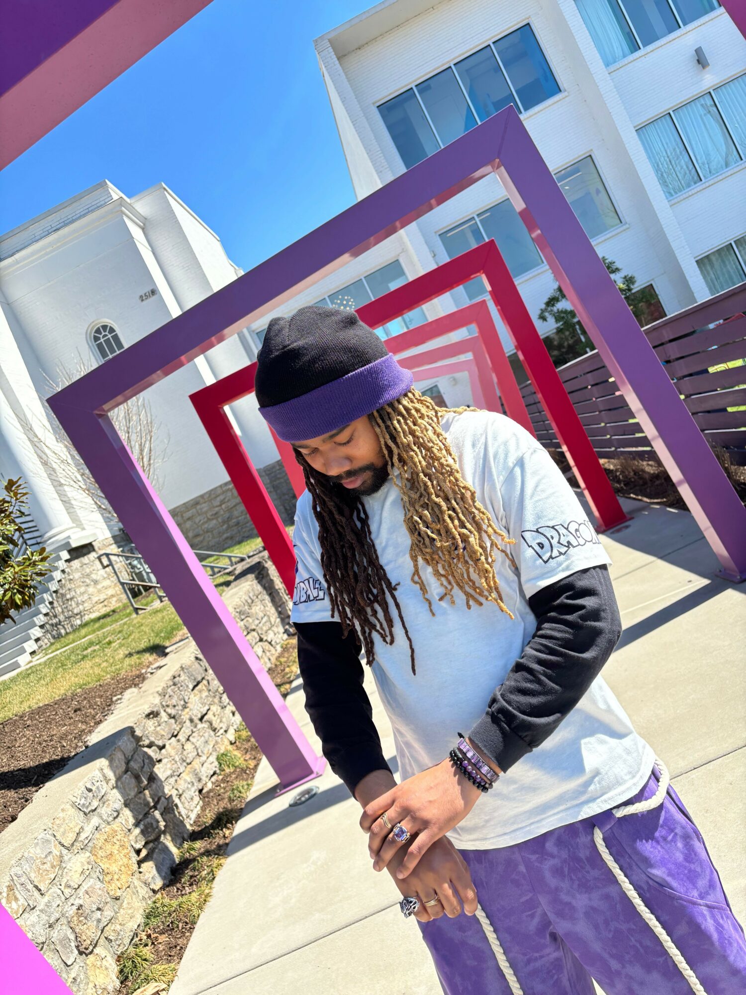 Person with long dreadlocks wearing a hat and jacket, standing outdoors near colorful geometric frames, holding a phone.
