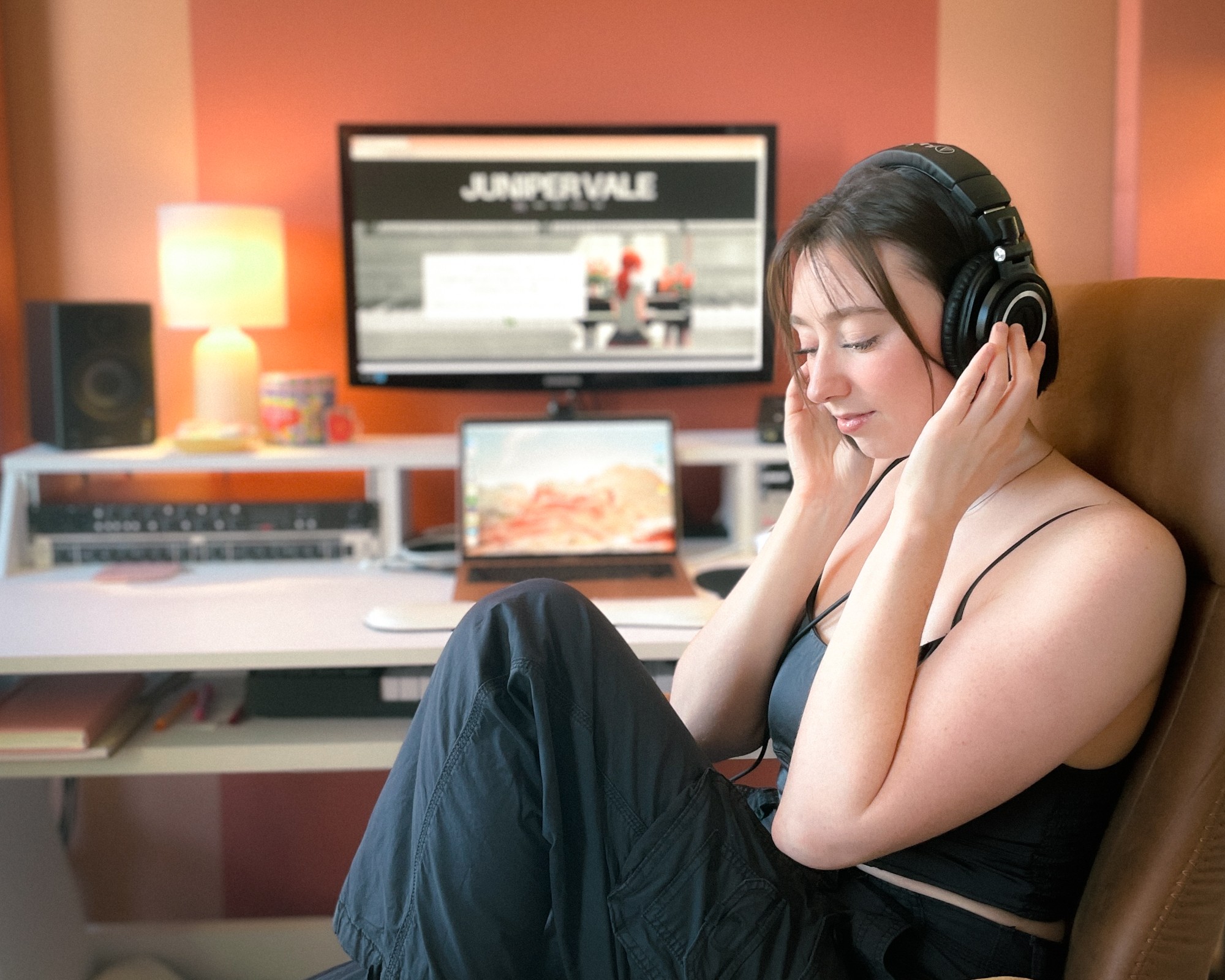 Young woman with headphones sitting in a room with a computer and monitor, eyes closed, relaxed, pink walls, warm lighting.