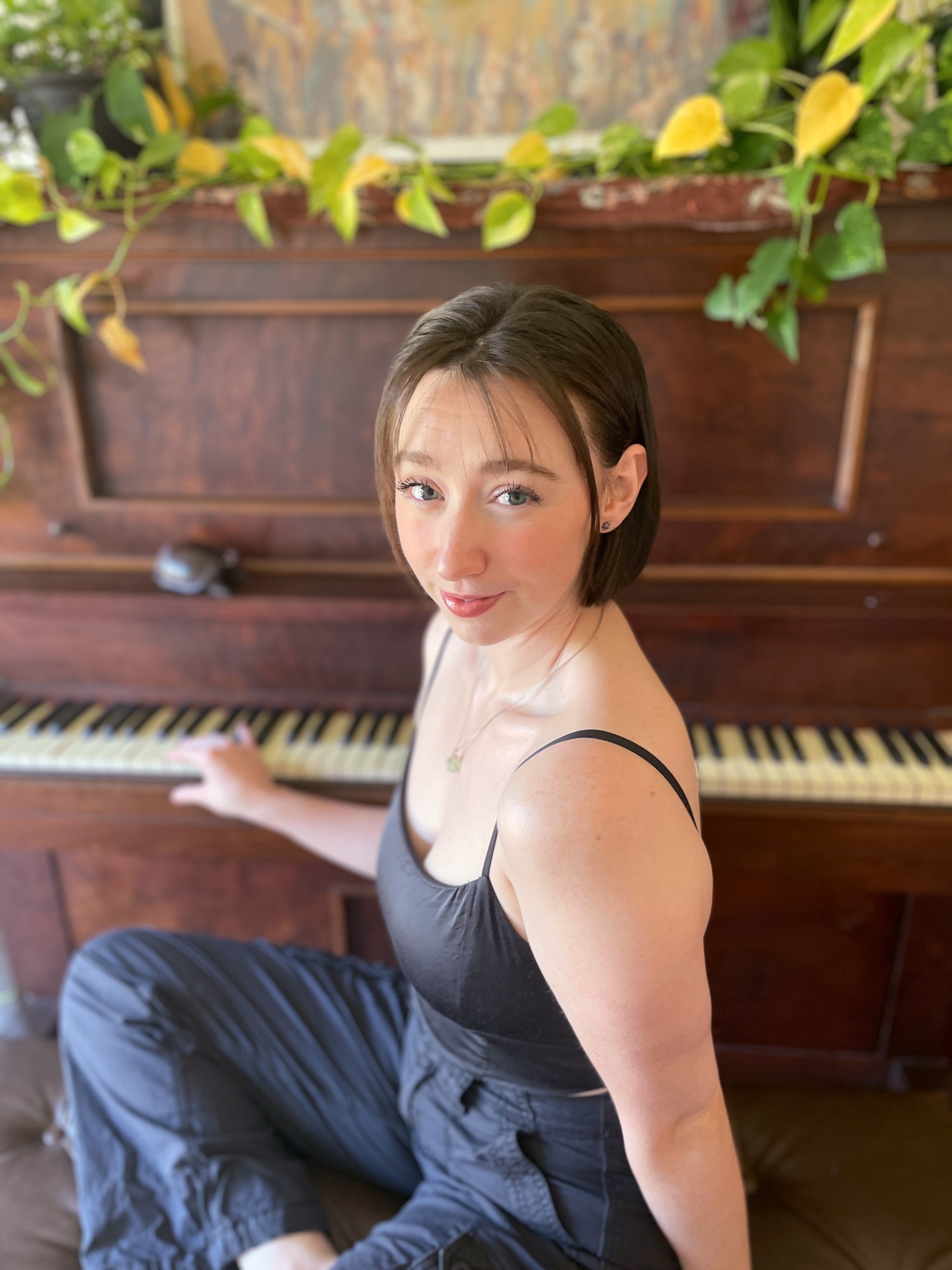 Young woman with short hair in a black dress sitting at a piano, looking at the camera, with plants above.