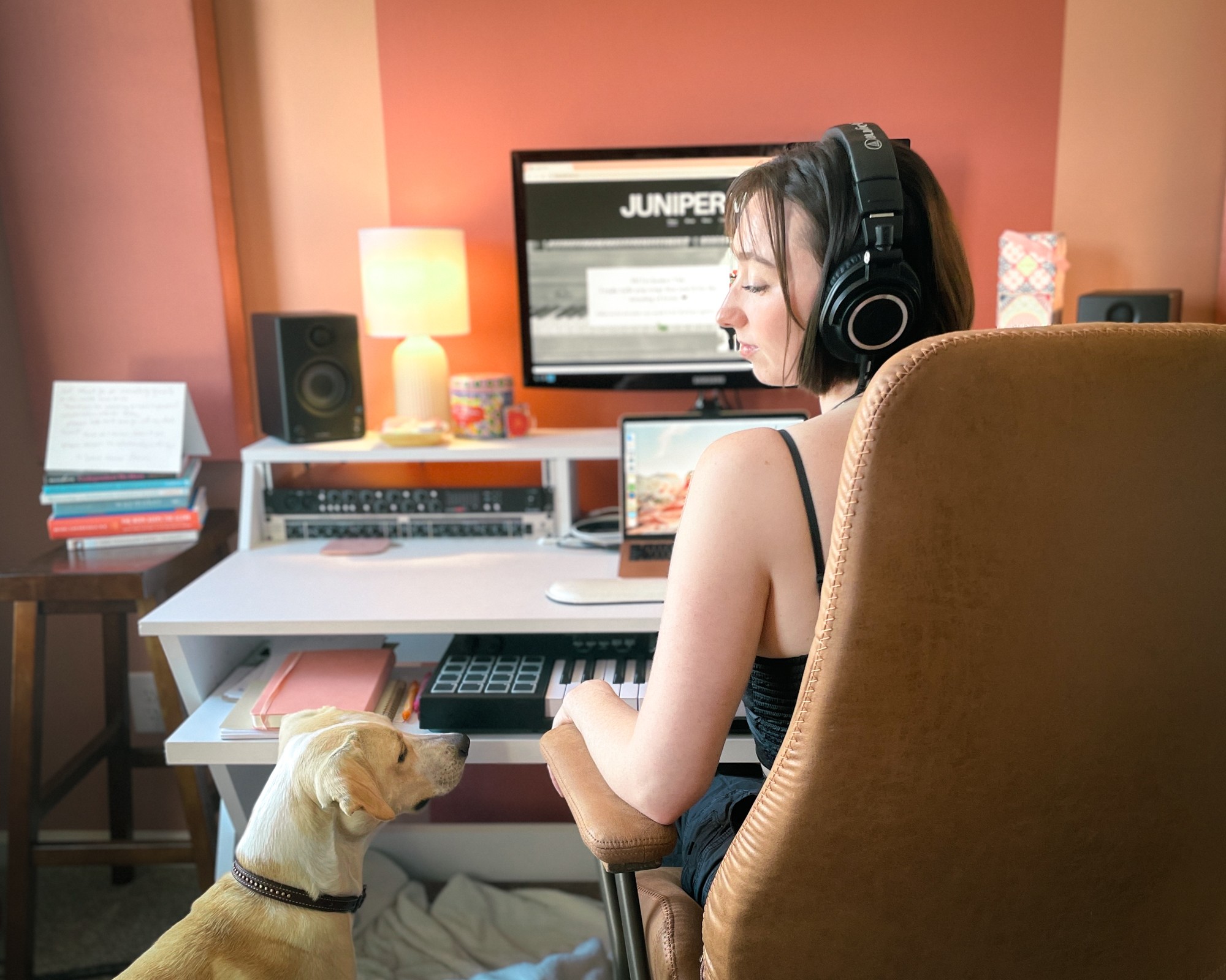 Woman wearing headphones working at a desk with a dog nearby, computer monitor in background, warm lighting.