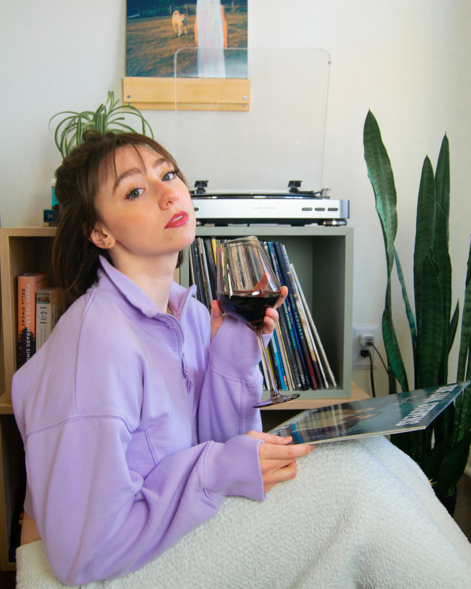 Young woman with short brown hair and purple sweatshirt holding a glass of red wine and a magazine, sitting indoors with plants and books.