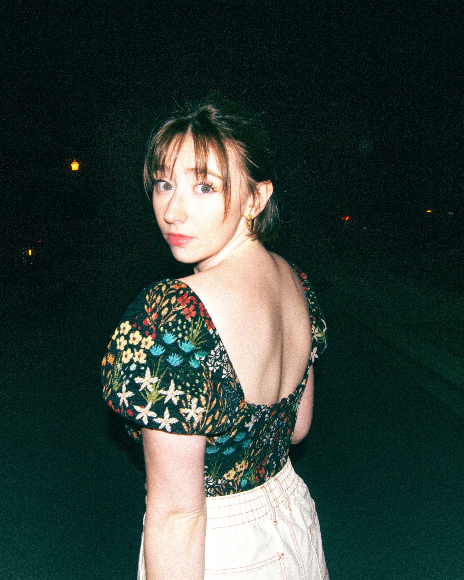 Young woman with shoulder-length hair and bangs, wearing a floral top with an open back, standing outdoors at night.