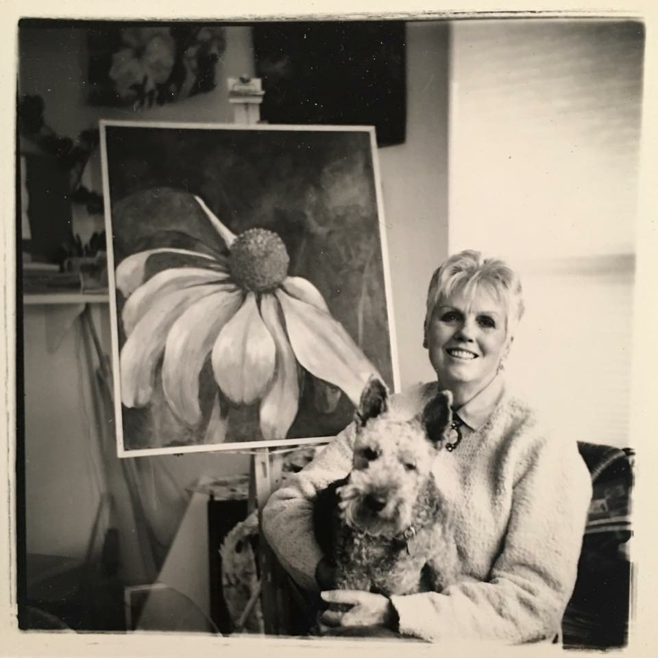Woman sitting with a dog in front of a large flower painting, smiling, in a room with window blinds.