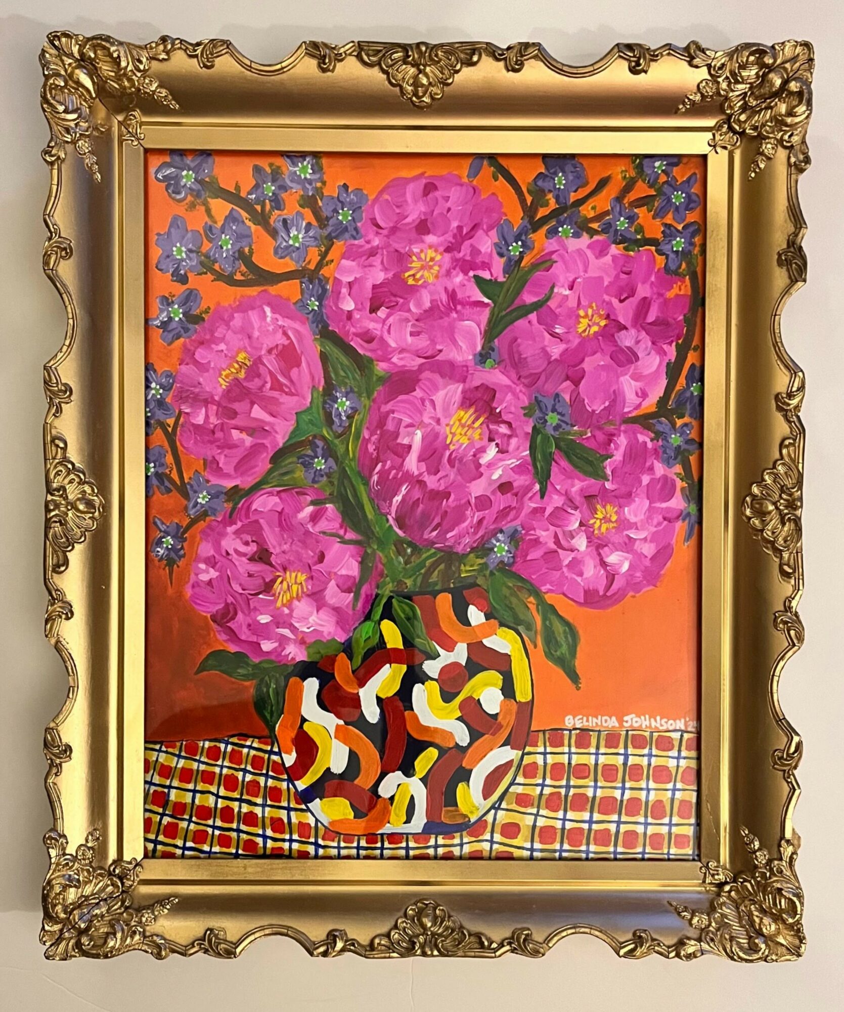 Vase with large pink flowers on a checkered tablecloth, framed in an ornate gold frame.