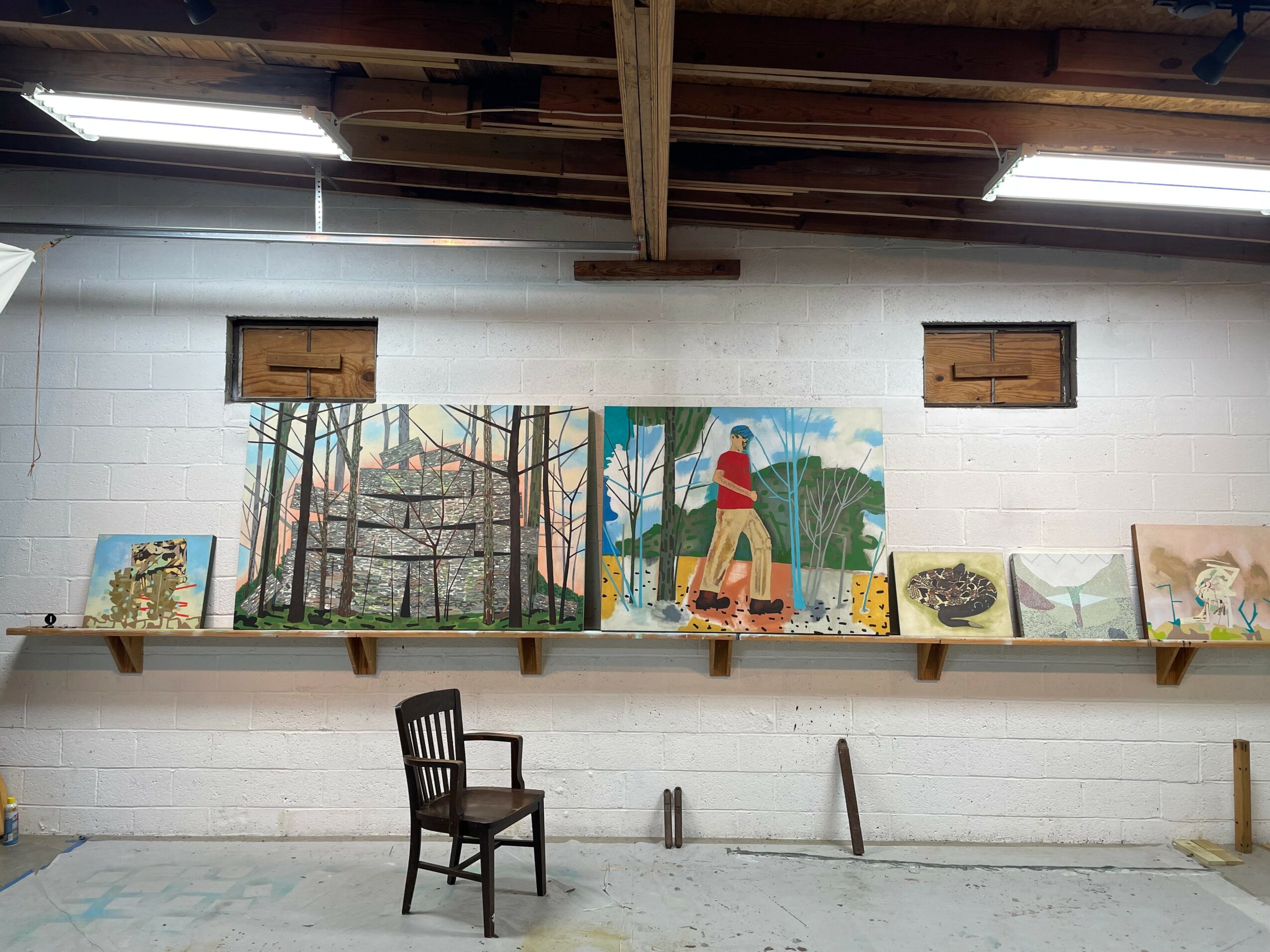 Paintings of trees and nature scenes on a white wall with a shelf below, a chair in front, and a wooden ceiling.