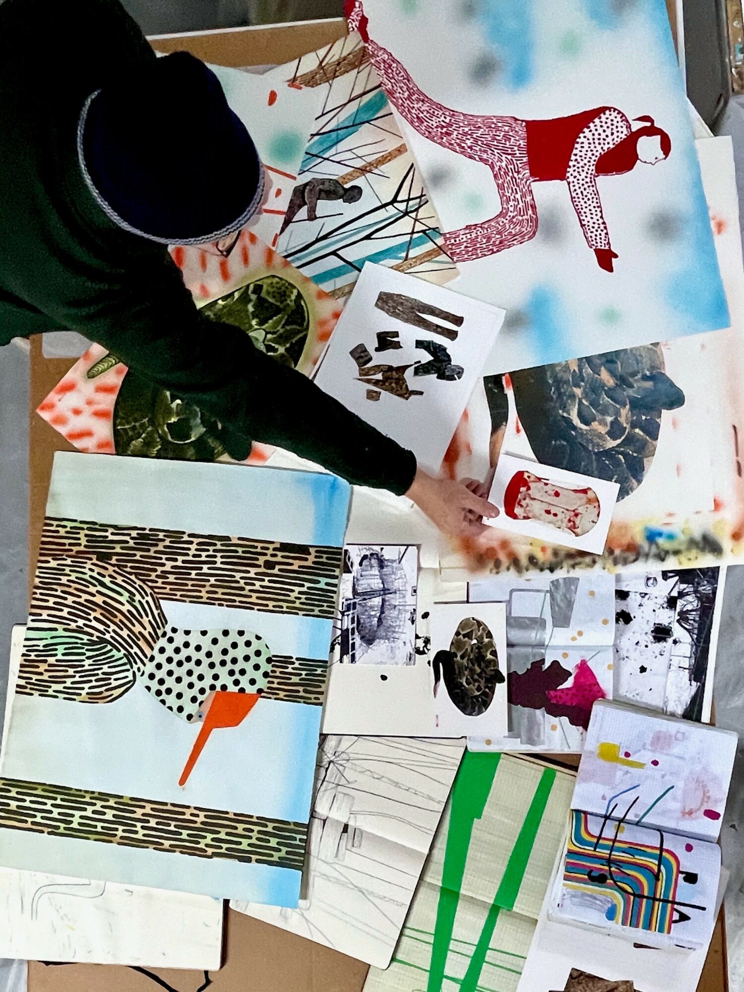 Person reaching over table with various art supplies, colorful paper, and artwork, viewed from above.