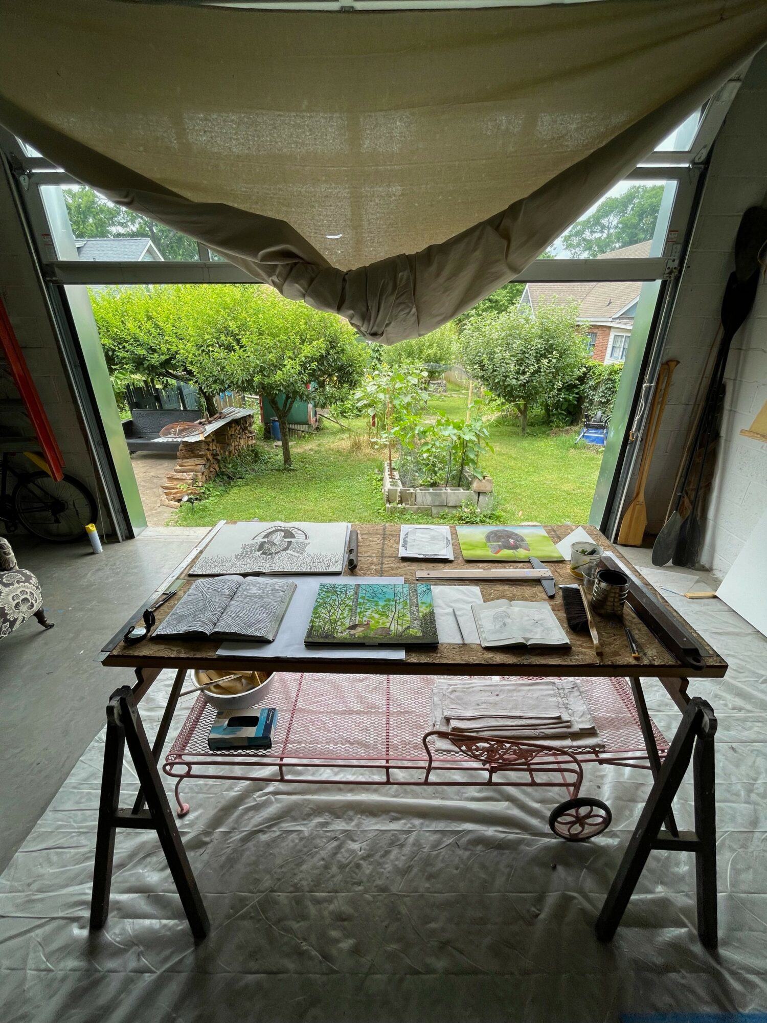 Table with books and papers in front of an open window showing a garden with trees and grass.