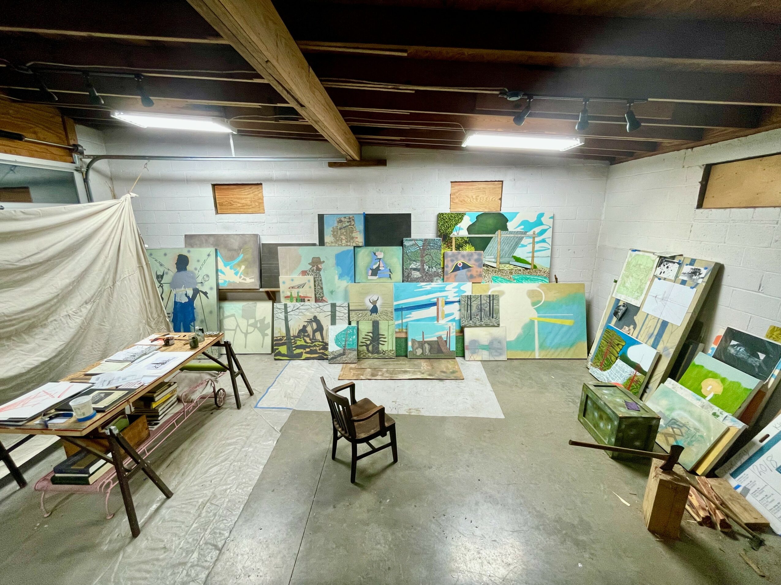 Art display with paintings and boards arranged in a room with wooden ceiling beams and concrete floor.