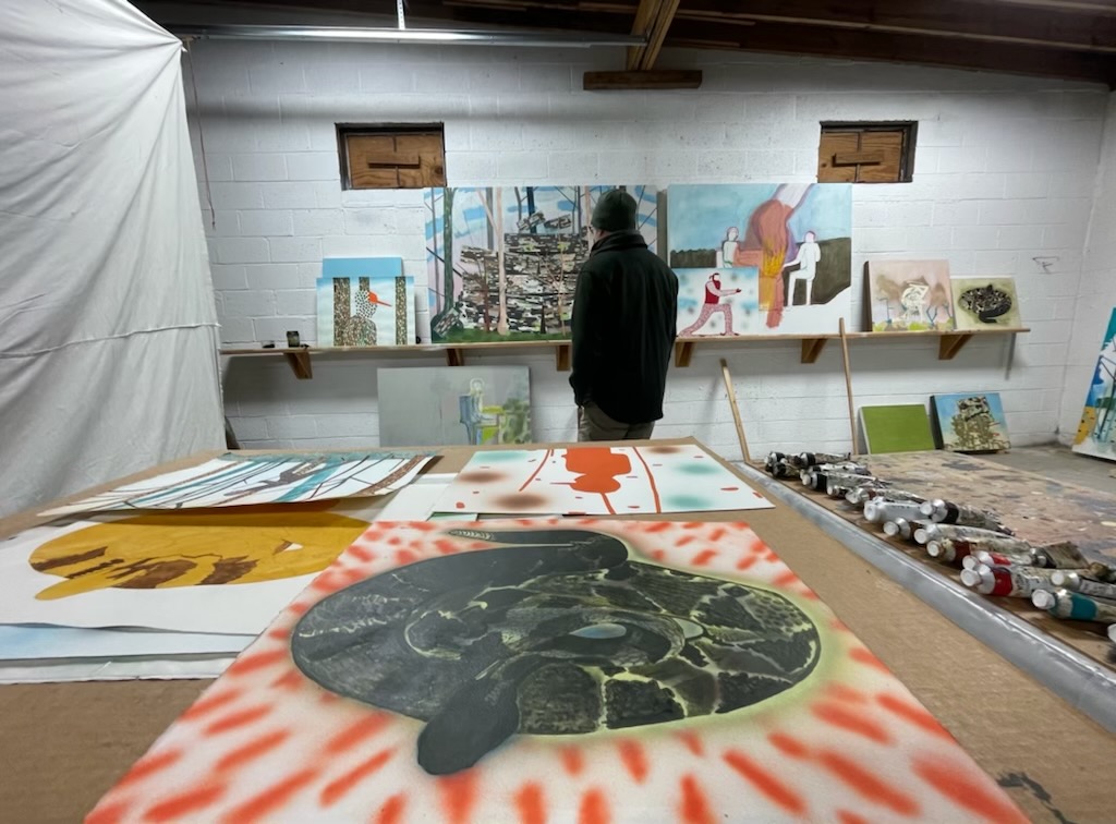 Person standing in an art studio with paintings and canvases on walls and tables, various artworks displayed around.
