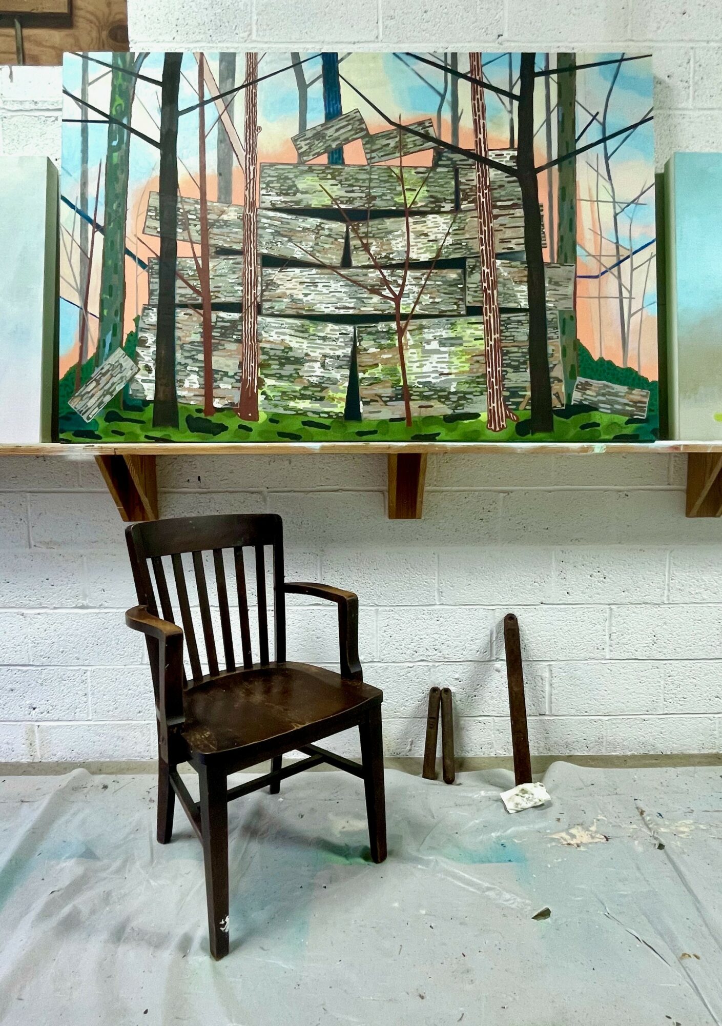 Painting of a forest scene with trees and a cityscape in the background, mounted on a wall above a wooden chair.