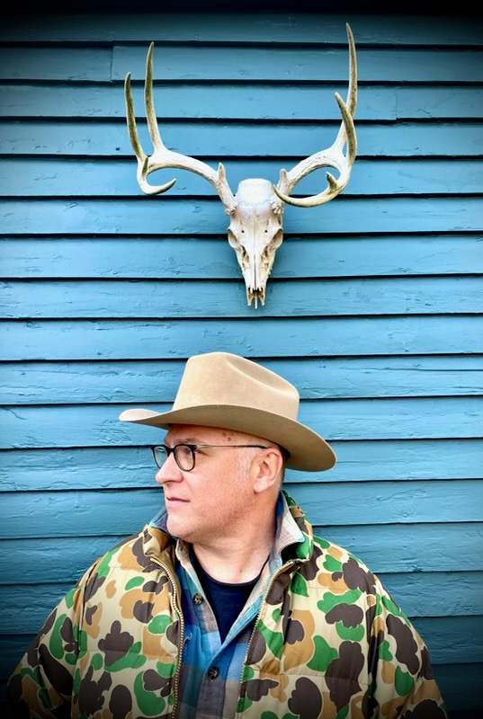 Man wearing a wide-brimmed hat and camouflage jacket stands in front of a blue wooden wall with a deer skull and antlers mounted above him.