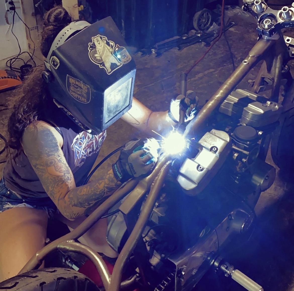 Person welding a motorcycle frame with protective gear, sparks flying, in a workshop setting.