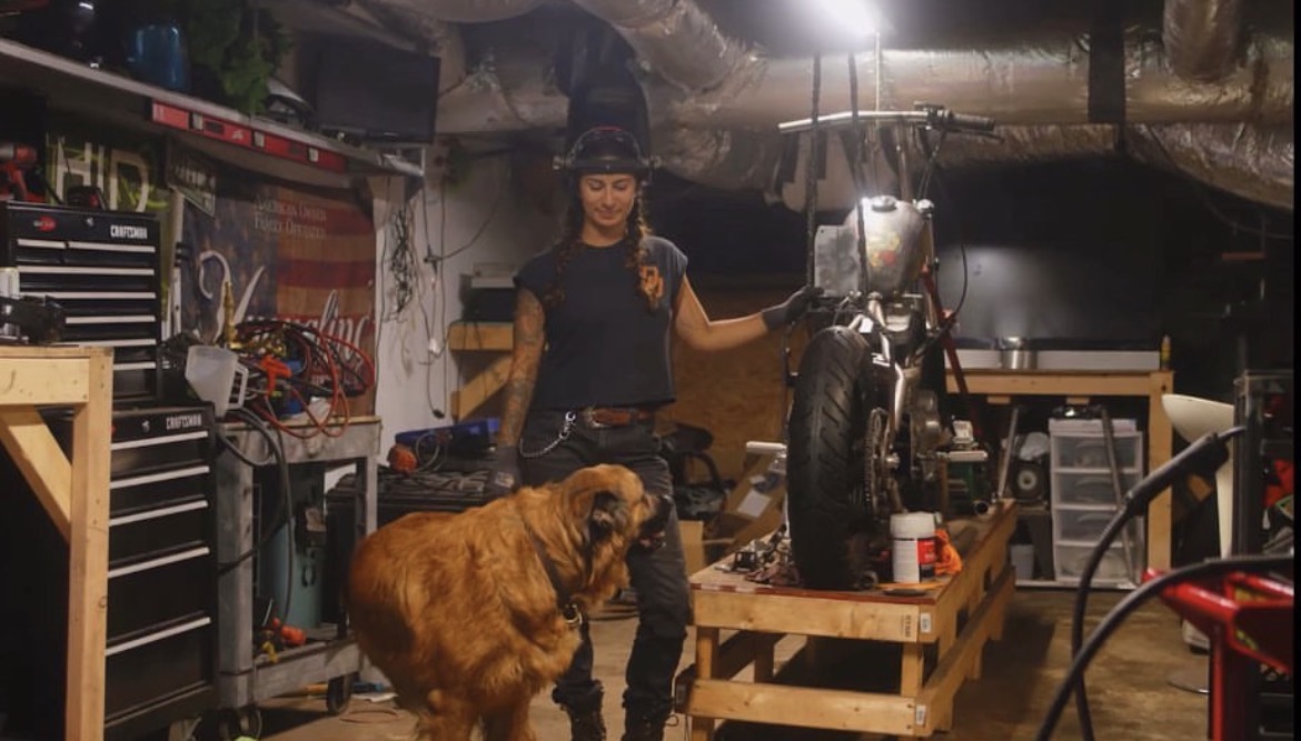 Person standing in a workshop with a large dog, surrounded by tools and equipment, holding a motorcycle handlebar.