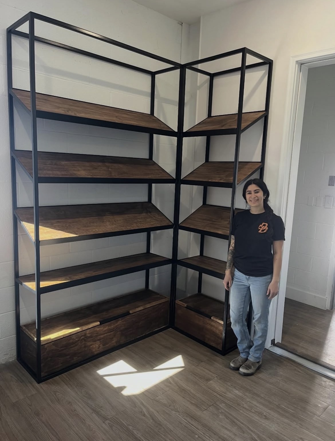 Person standing next to a large black metal and wood shelving unit in a room with wooden floor and white walls.