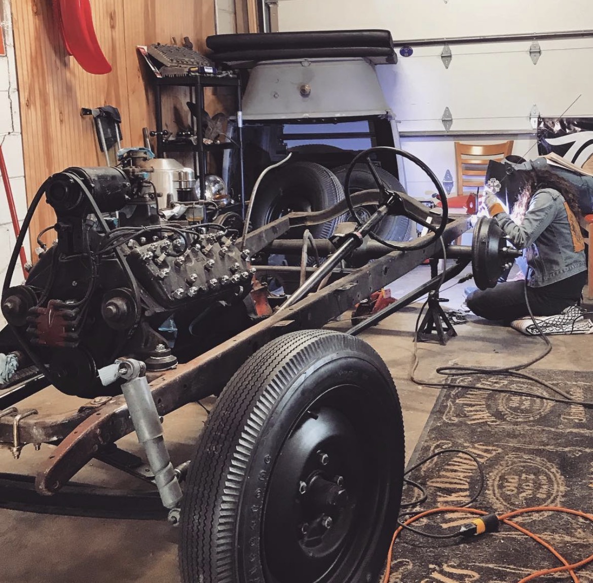 Person working on a car chassis with engine in a garage, tools and equipment around, wooden wall in background.
