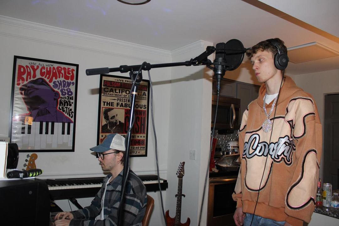Two young men in a music studio, one playing keyboard and the other singing into a microphone, with posters on the wall.