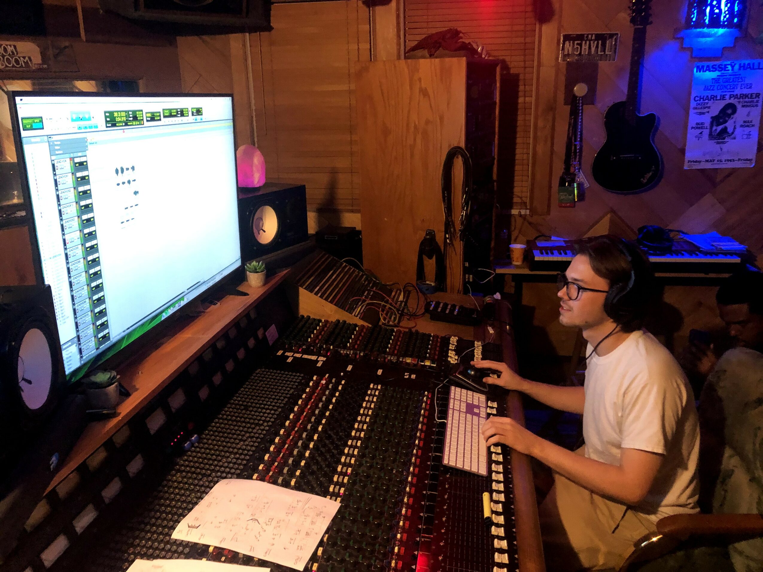 Person working at a music mixing console with a large screen, speakers, and musical instruments in a recording studio.