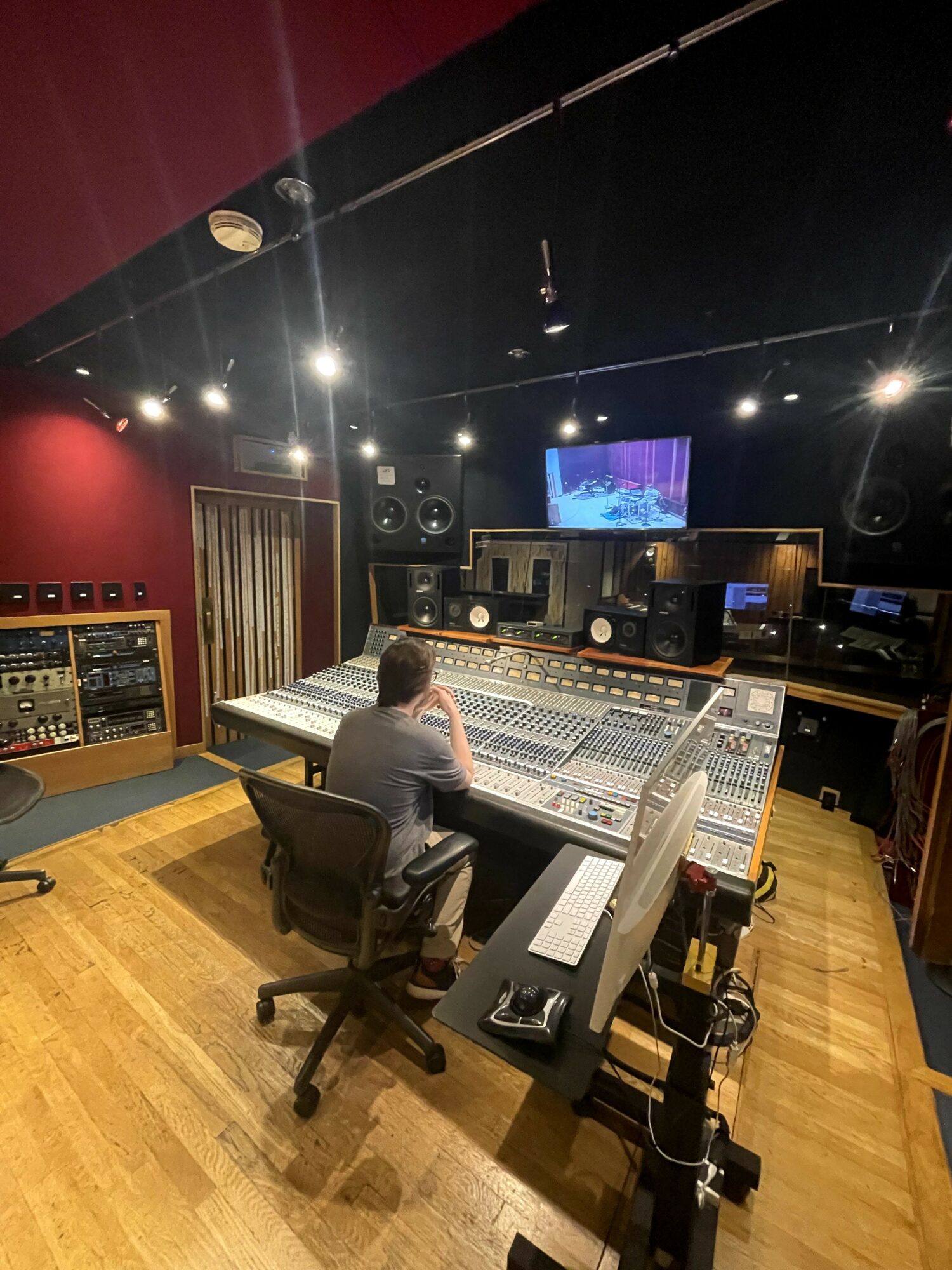 Person sitting at a large mixing console in a recording studio with equipment, speakers, and a TV screen on the wall.
