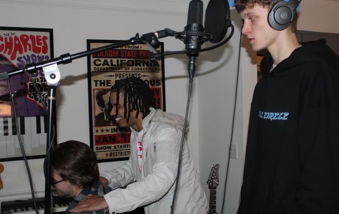 Two young men in a recording studio, one with headphones singing into a microphone, the other working on a keyboard.