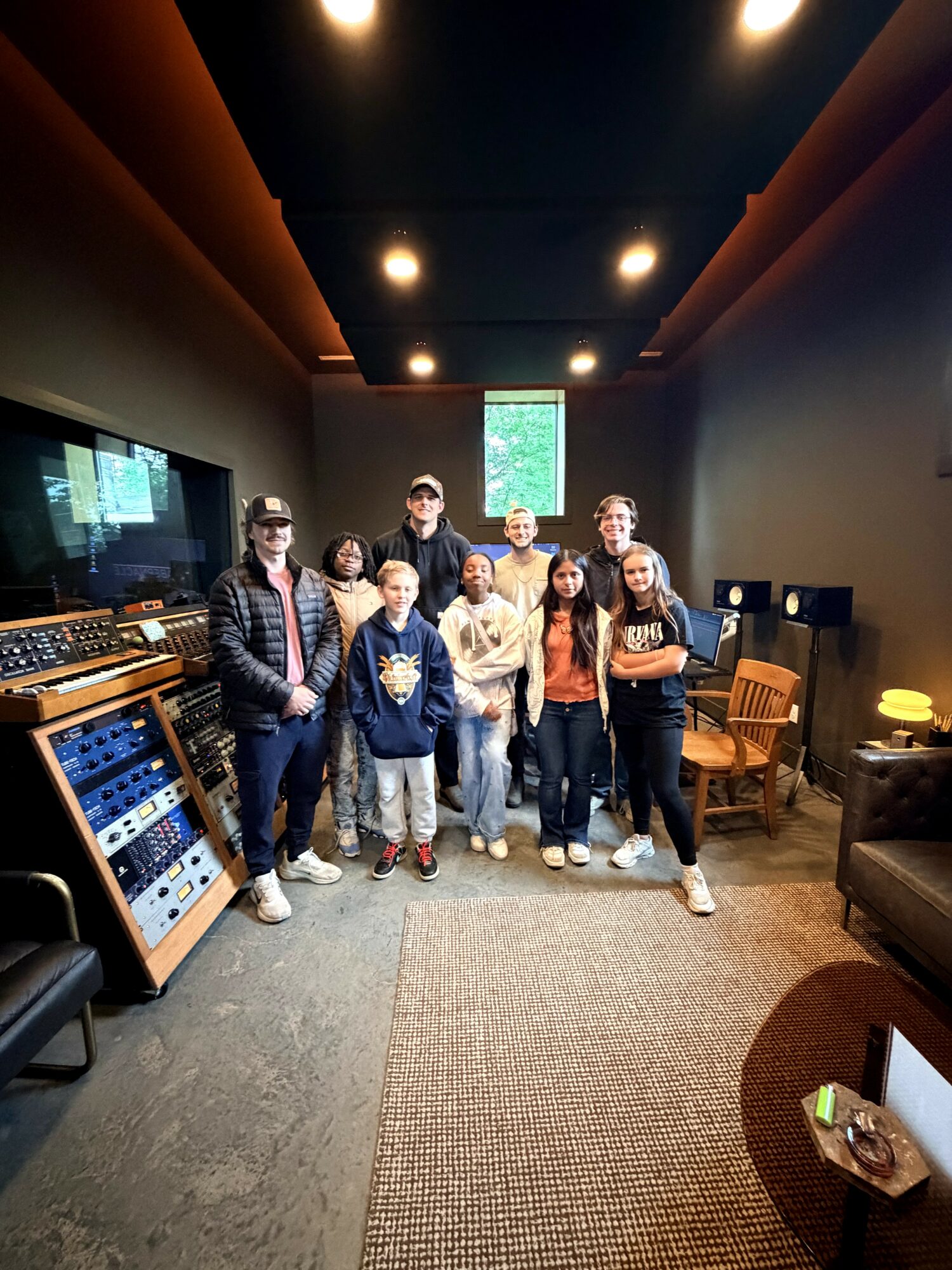 Group of nine people standing in a recording studio with equipment and chairs, some children and adults, facing the camera.