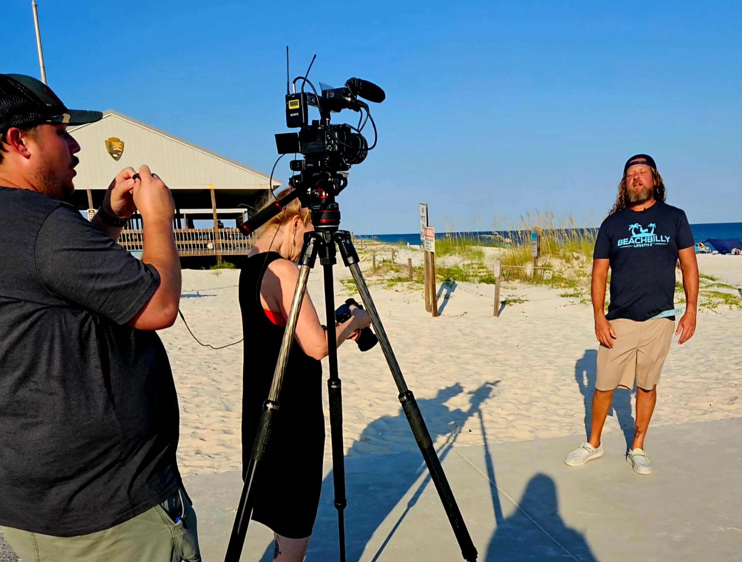 People filming a person on a sandy beach with a camera and tripod, blue sky, and ocean in background.