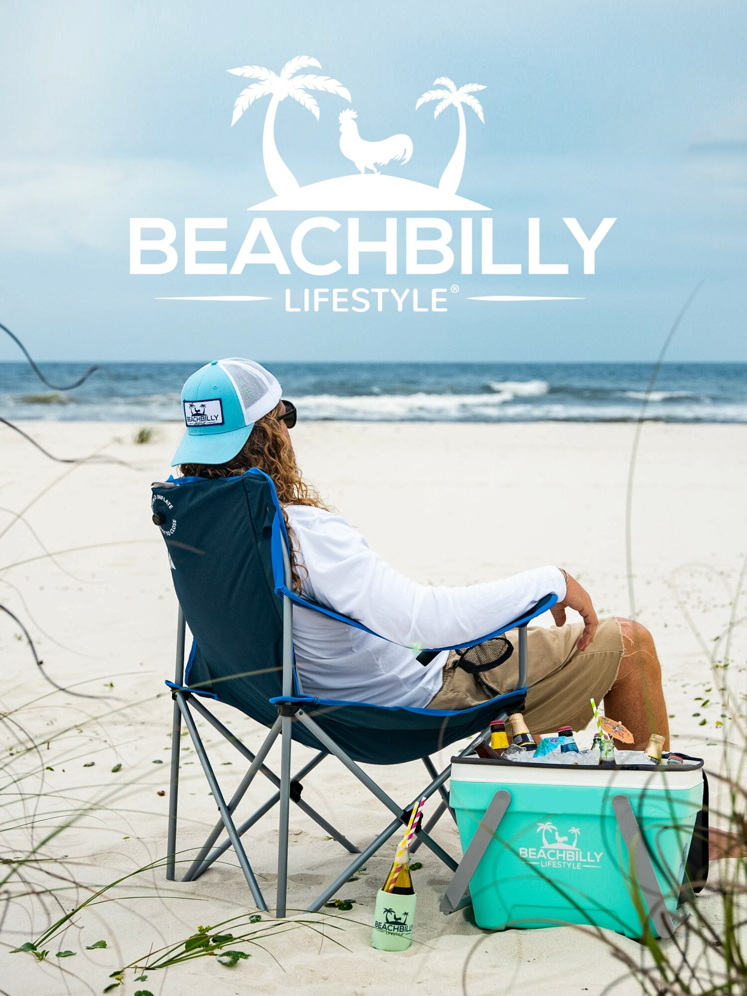 Person sitting on a beach chair with a cooler, fishing rod, and beach accessories, facing the ocean under a clear sky.