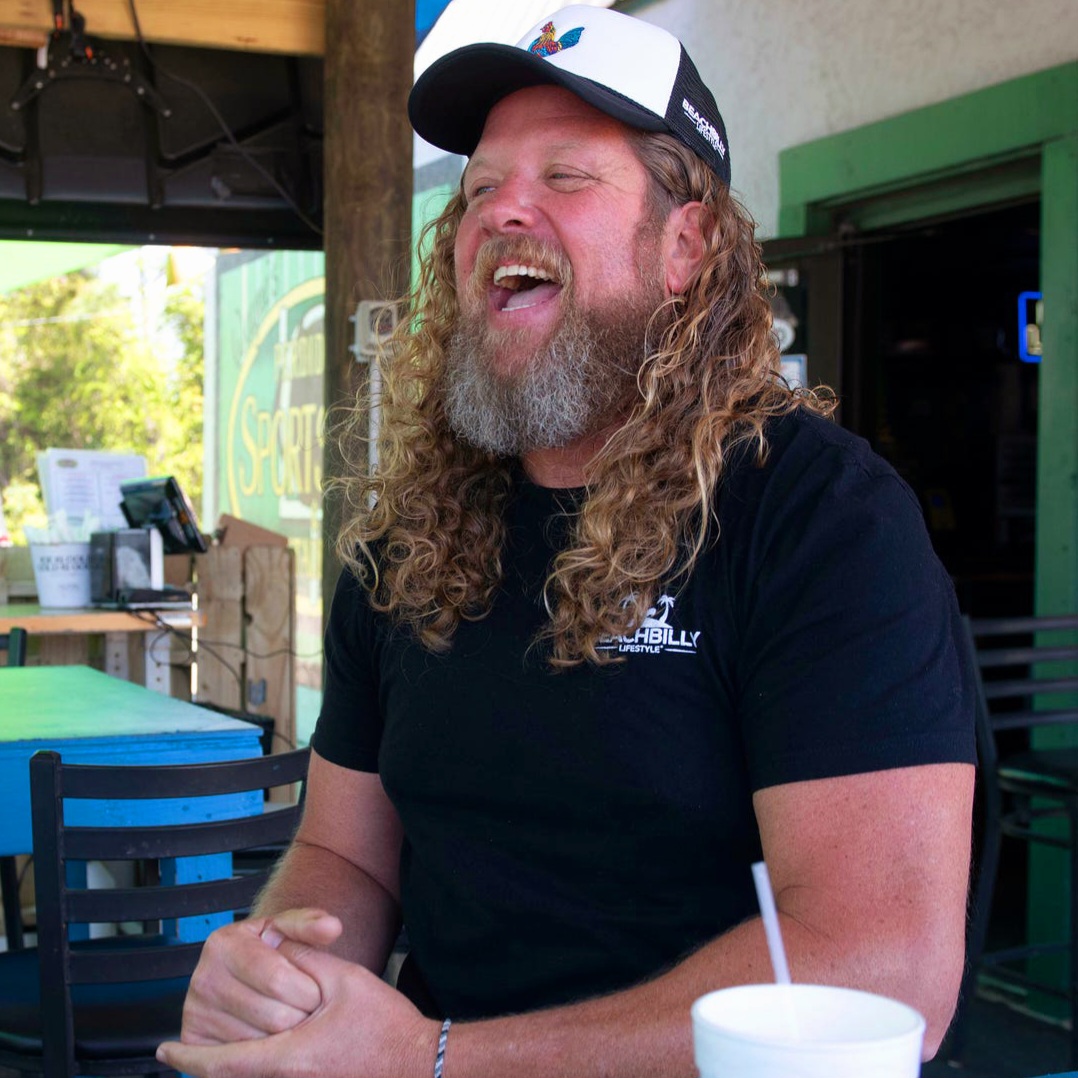 Man with long curly hair and beard laughing outdoors, wearing a black shirt and a white cap, sitting at a table.
