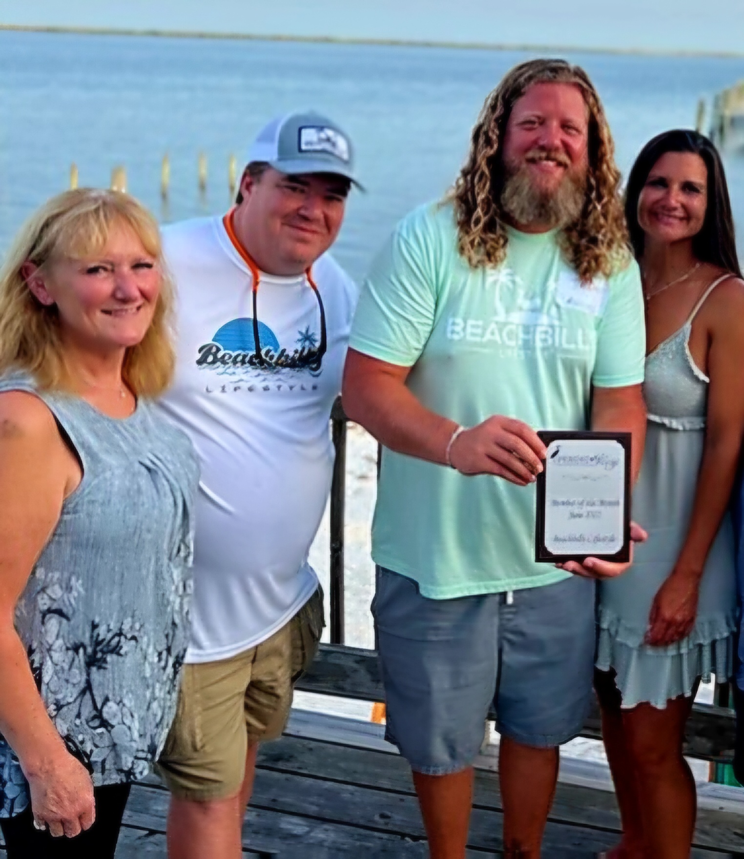 Four people stand outdoors near water, smiling, with a man holding a plaque. Background shows water and a dock.
