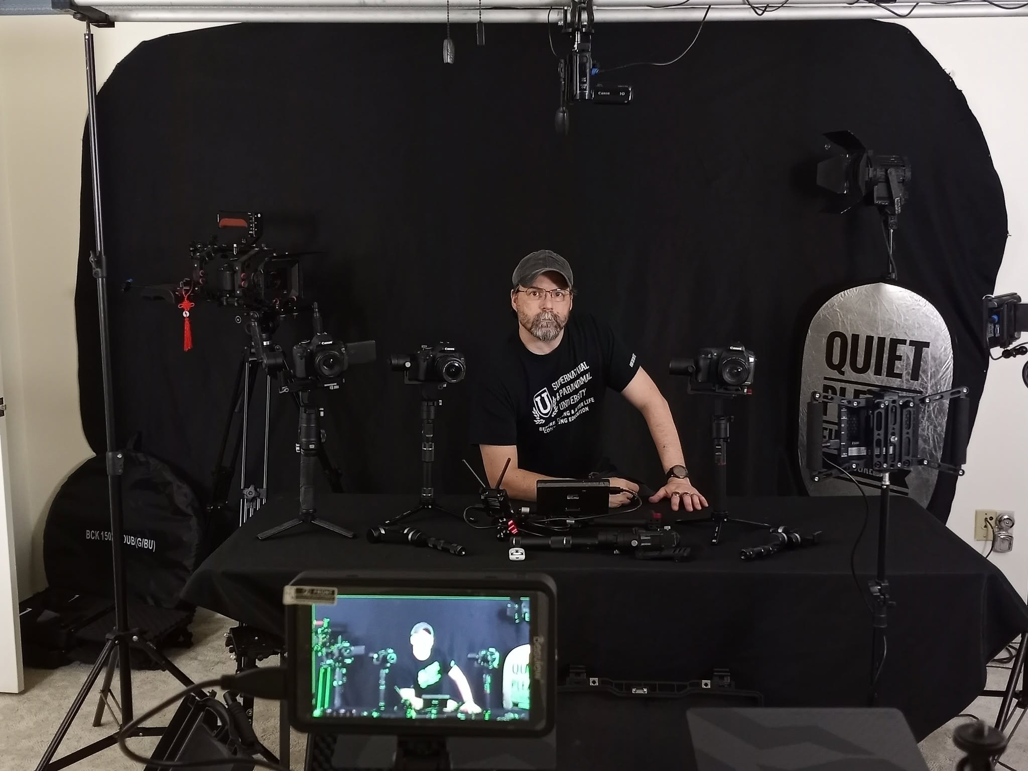 Man with a beard and glasses sitting at a table with camera equipment, in front of a black backdrop, with a sign reading 'QUIET'.
