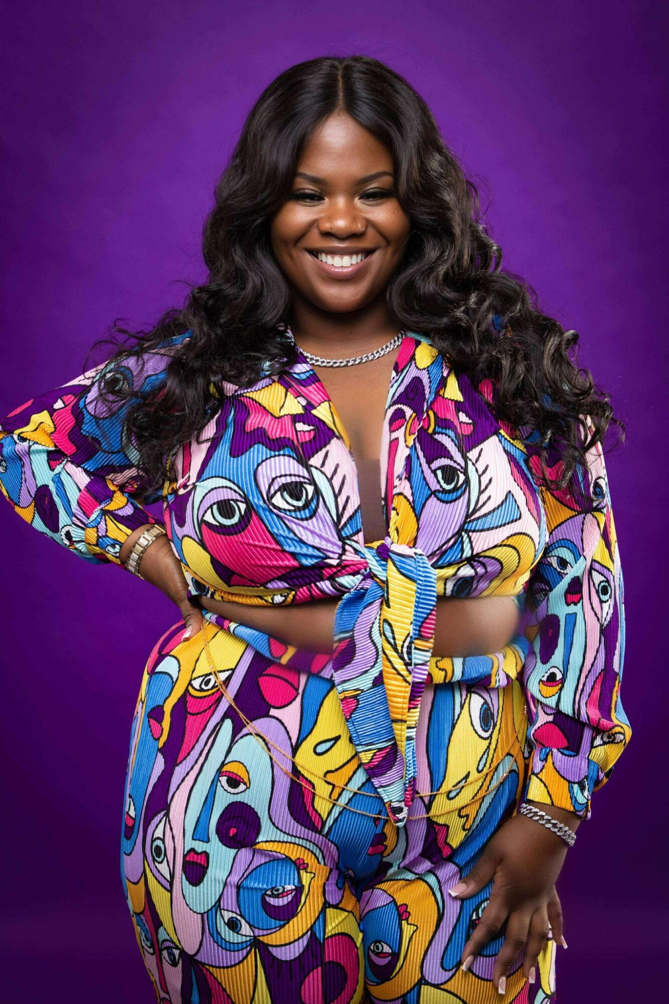 Smiling woman with long curly hair wearing a colorful patterned dress and a pearl necklace, standing against a purple background.
