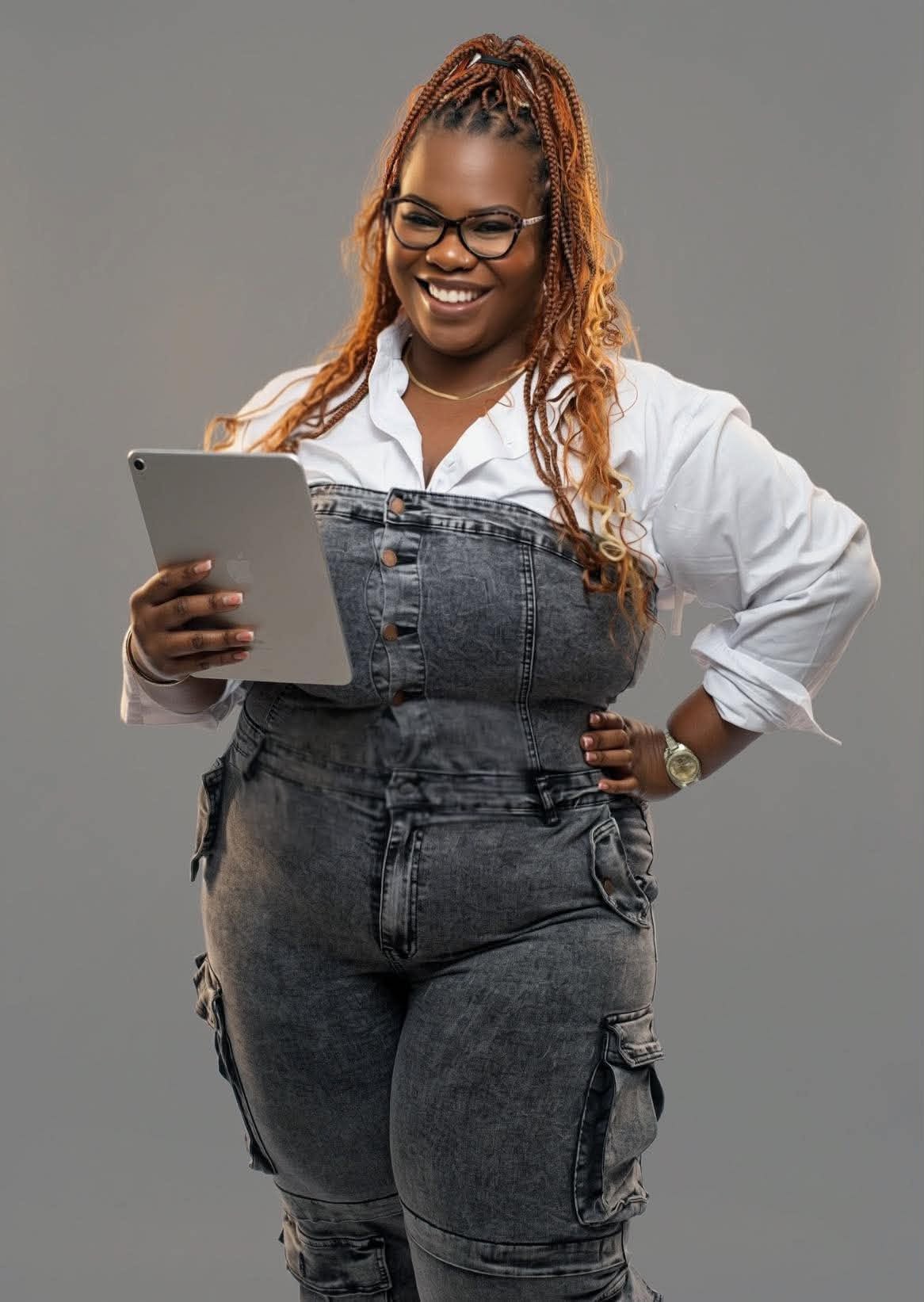 Smiling woman with glasses holding a tablet, wearing a white shirt and denim overalls, standing against a gray background.