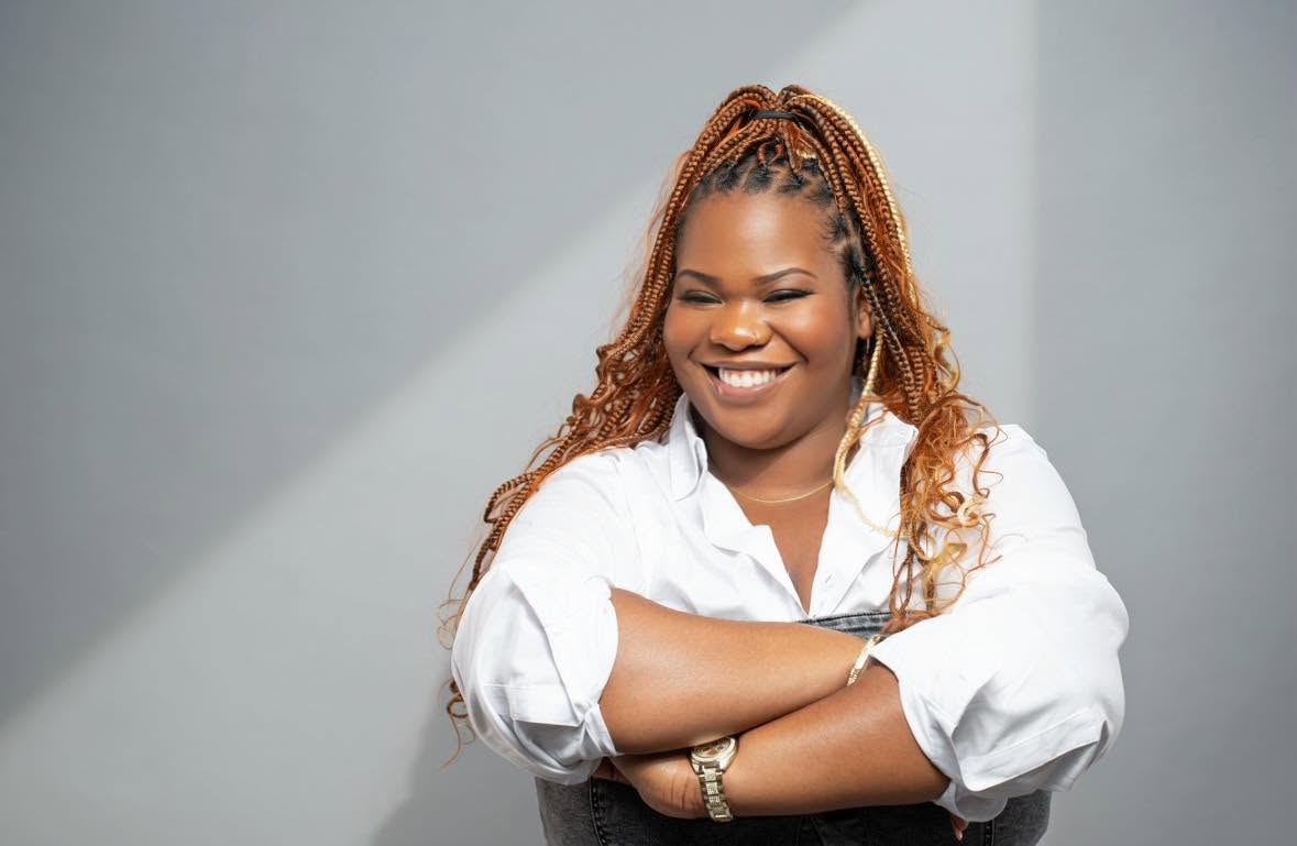 Woman with braided hair smiling, arms crossed, wearing a white shirt, standing against a plain background.