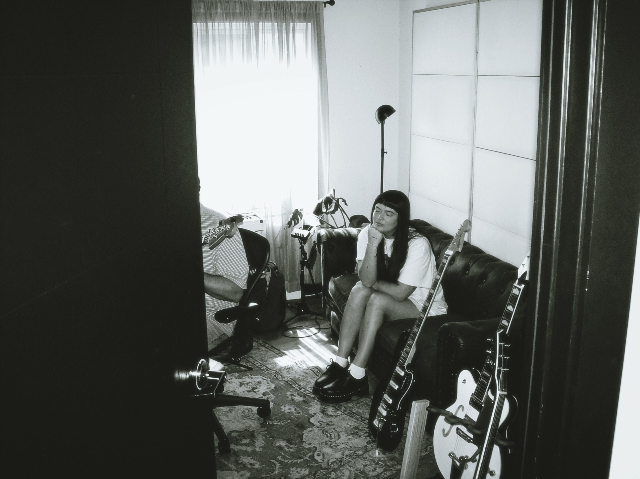 A girl sitting on a sofa in a room with musical instruments, a window with curtains, and a lamp.