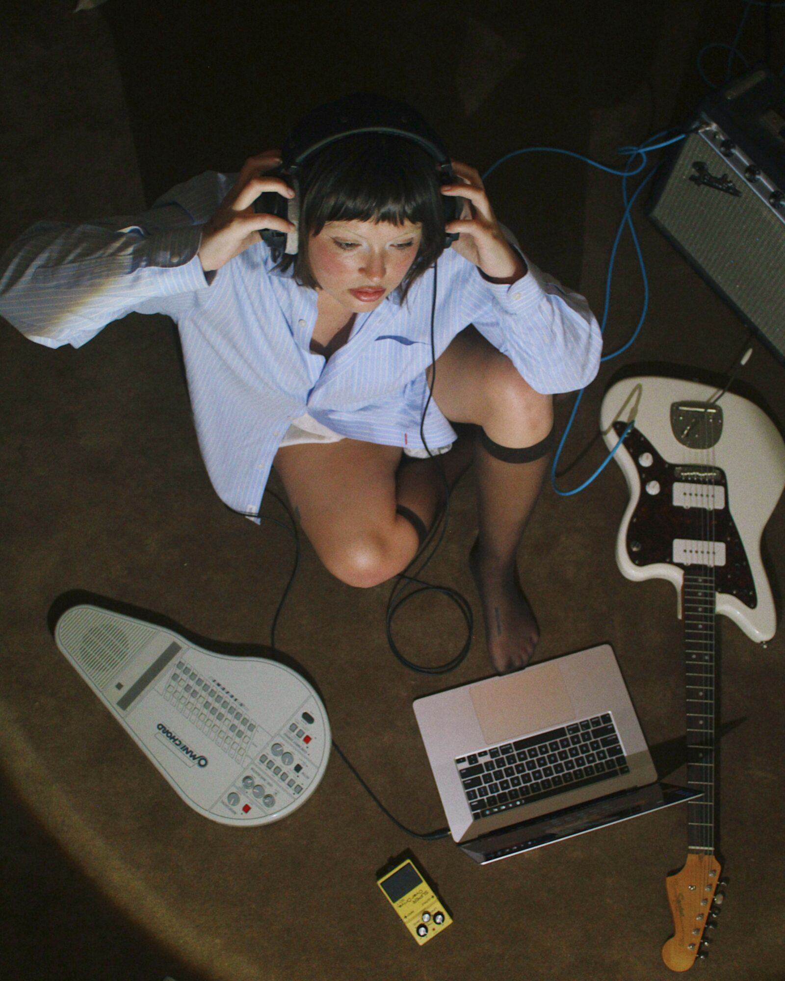 Person sitting on floor with headphones, surrounded by musical equipment, guitar, laptop, and electronic device, viewed from above.