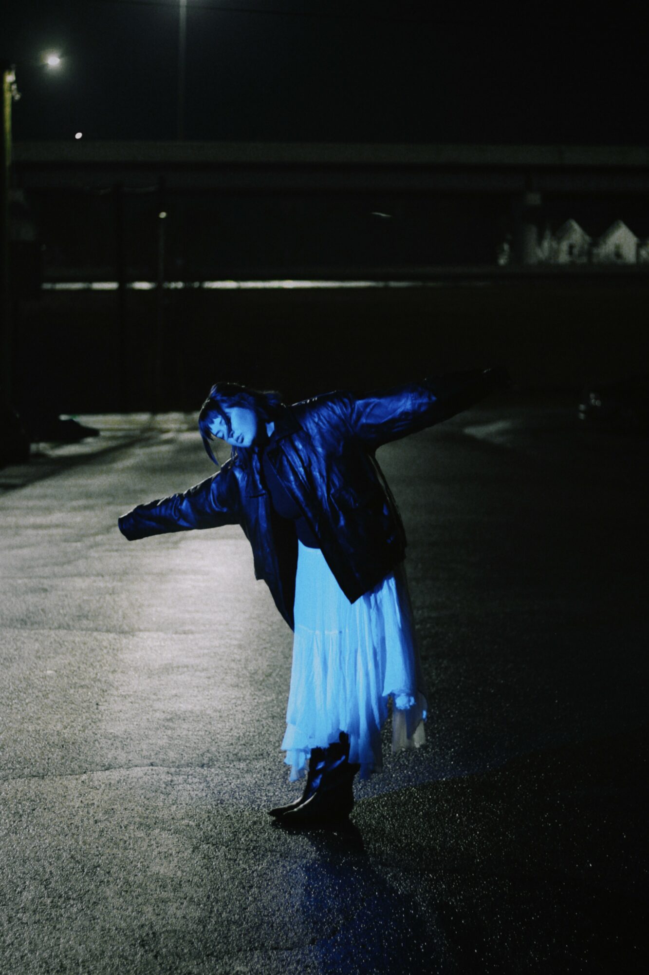 Person in a blue jacket and white dress ice skating outdoors at night, arms outstretched, on a dark surface.