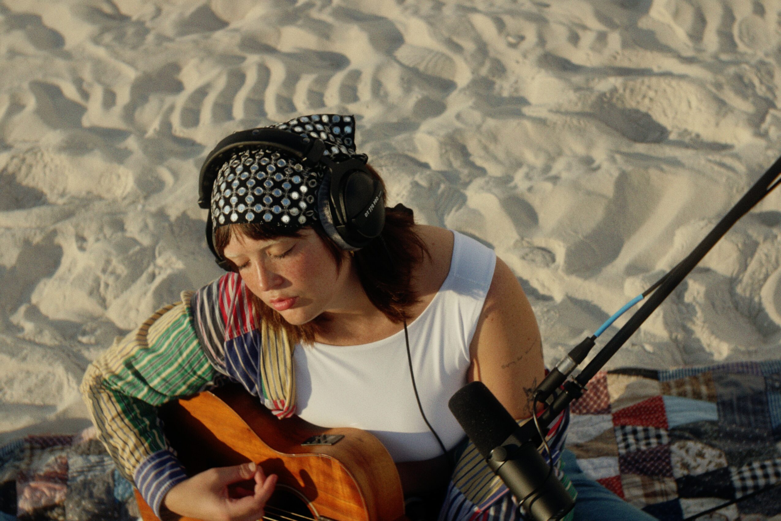 Woman wearing headphones playing guitar on sandy beach, with a patchwork blanket nearby.