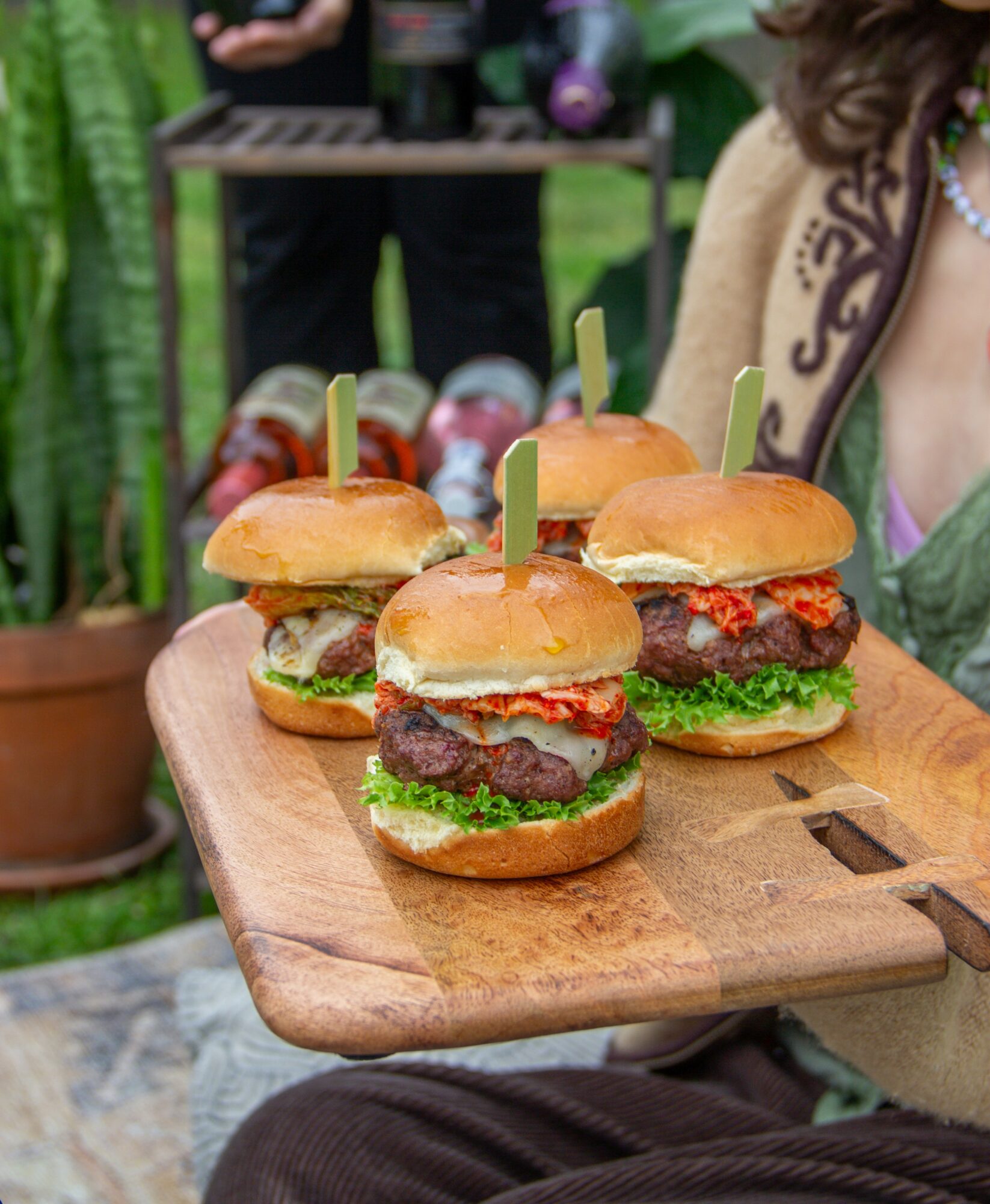 Four mini burgers with lettuce, tomato, and meat patties on a wooden serving board, outdoors with plants and people in background.
