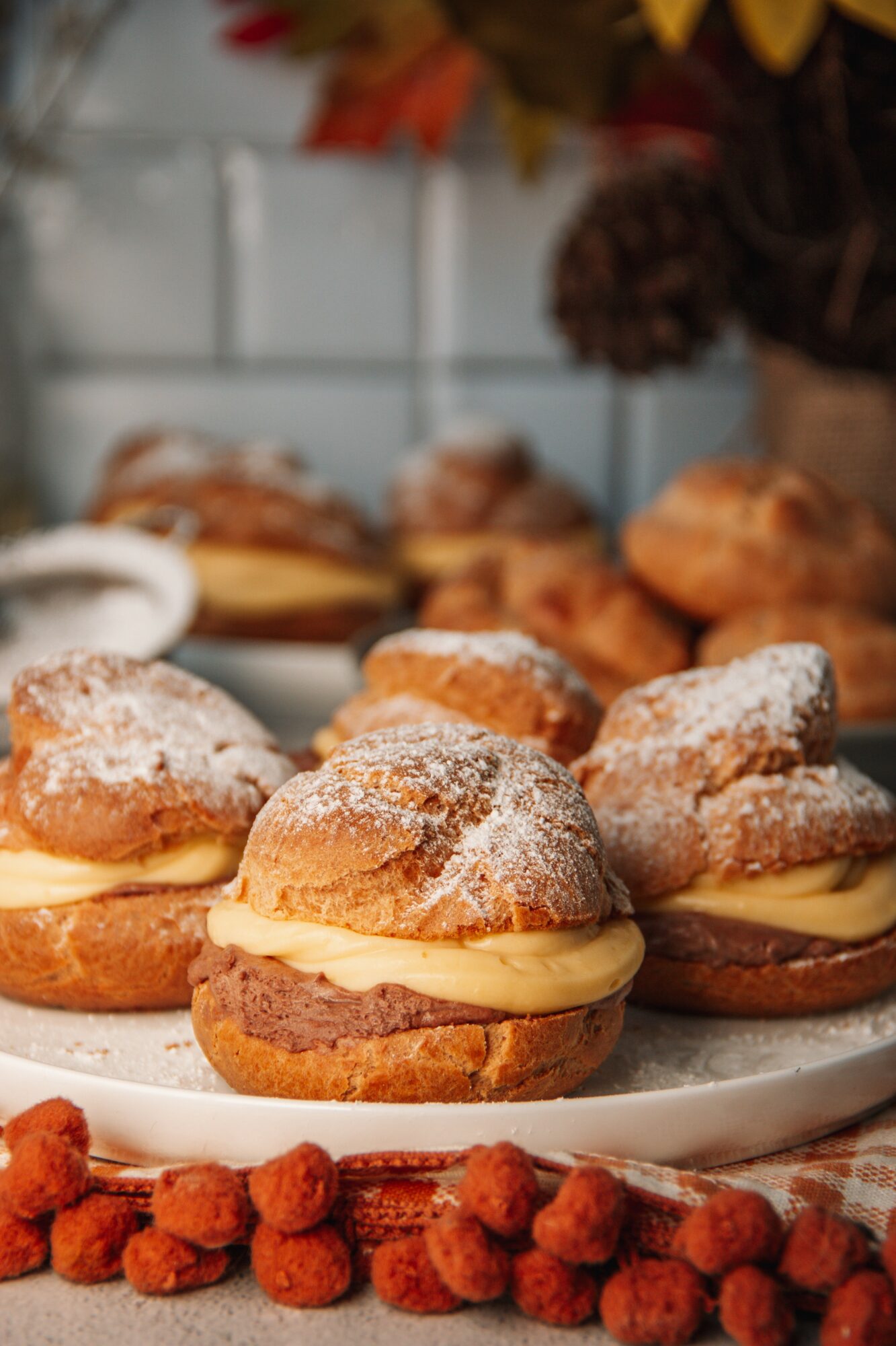 Cream-filled pastries dusted with powdered sugar on a white plate, with a blurred background of more baked goods and flowers.