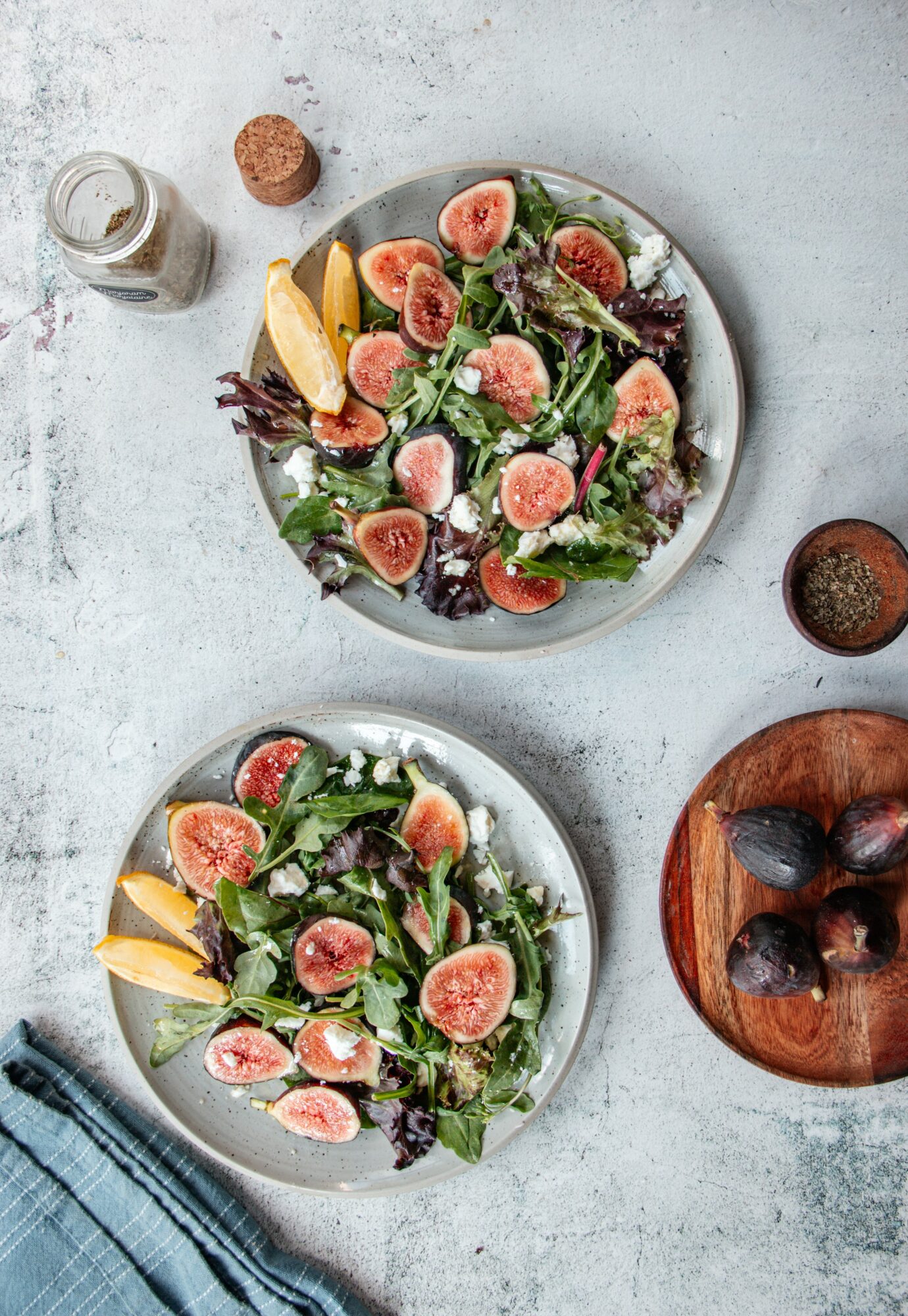 Two plates of salad with figs, lemon slices, and greens on a light surface, with a jar, cork, and wooden bowl nearby.