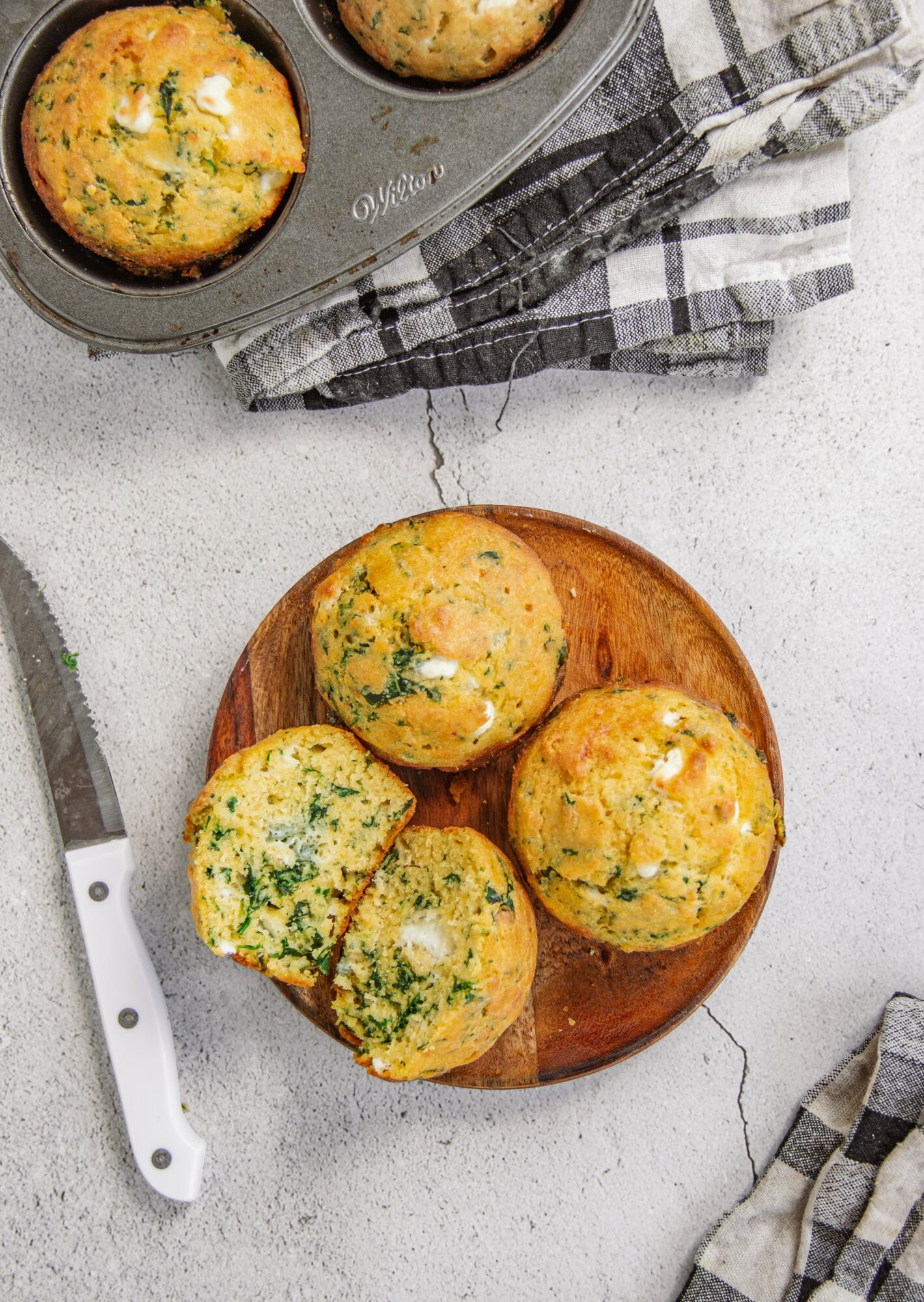 Four muffins on a round wooden plate, with a knife and checkered cloth nearby, on a white surface.