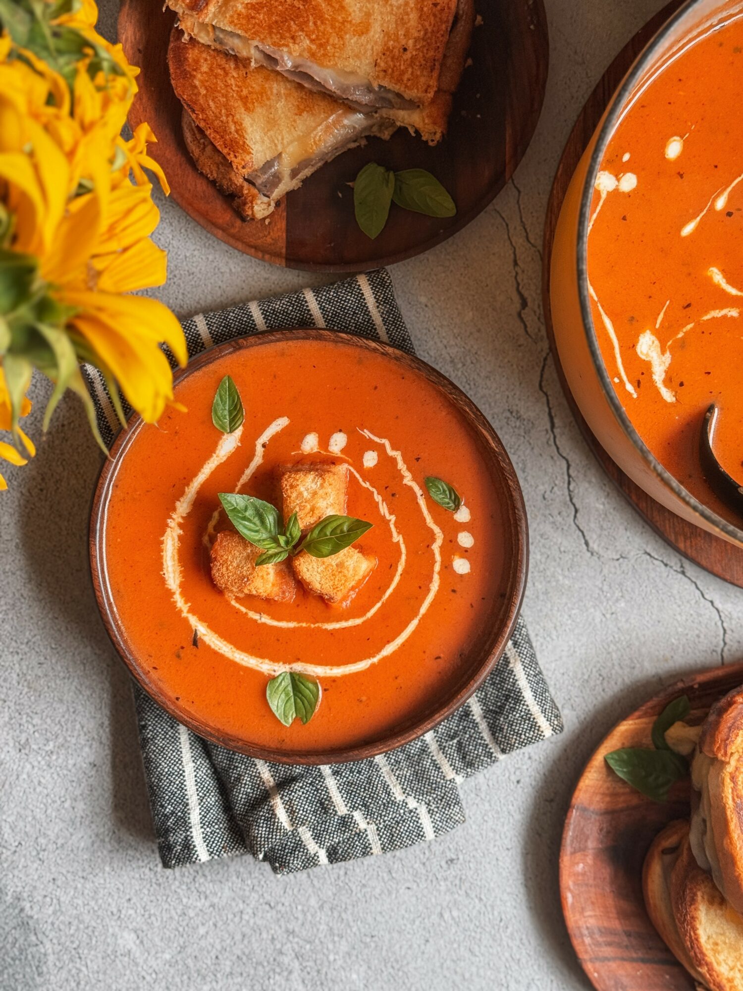 Bowl of tomato soup garnished with basil leaves and cream swirl, with bread on a plate nearby, on a striped cloth.
