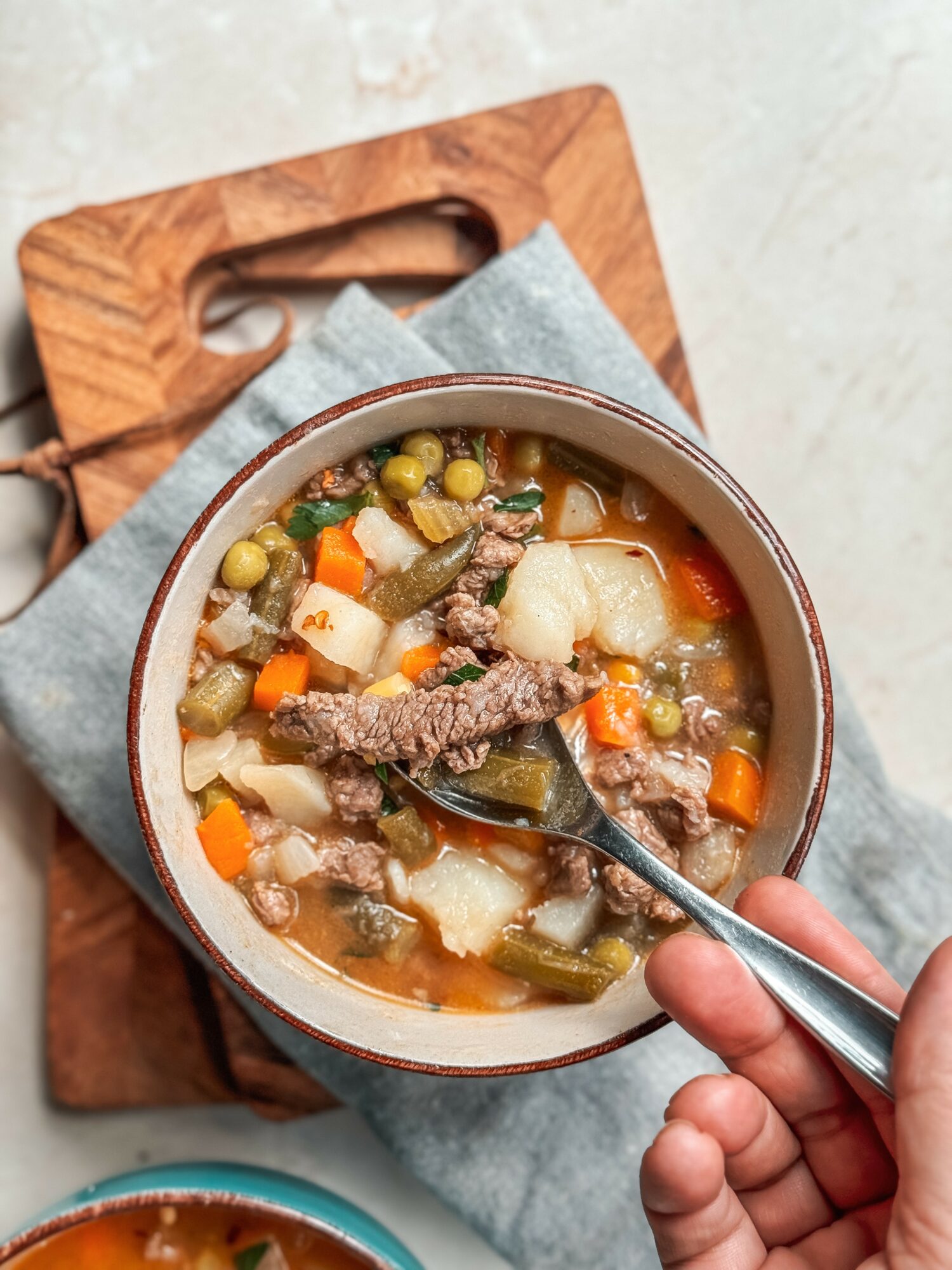 Bowl of vegetable and meat soup with a spoon, on a gray cloth, on a wooden tray, viewed from above.