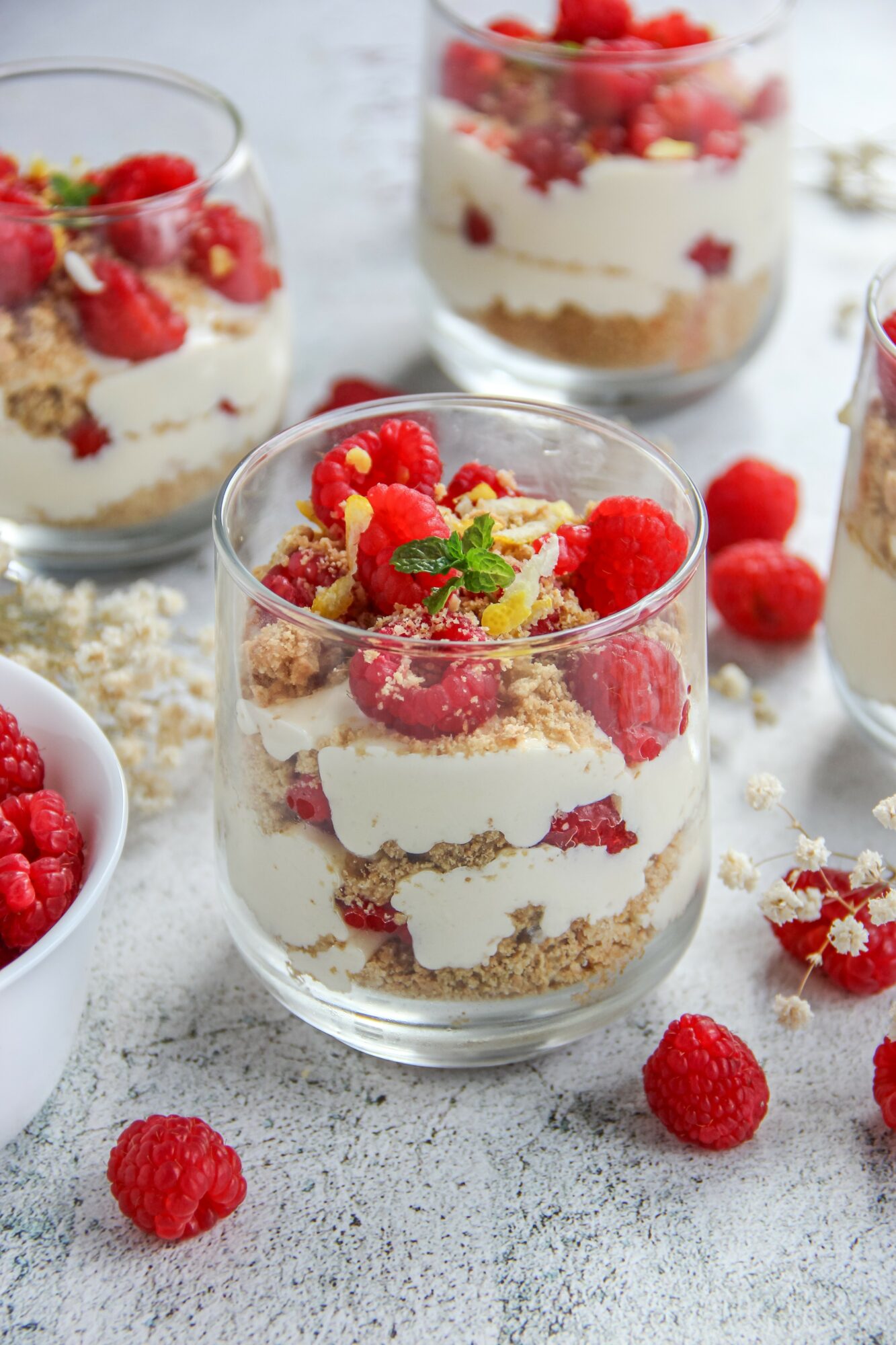 Layered dessert with cream, raspberries, and crumbs in a glass, garnished with berries and mint, on a white surface.
