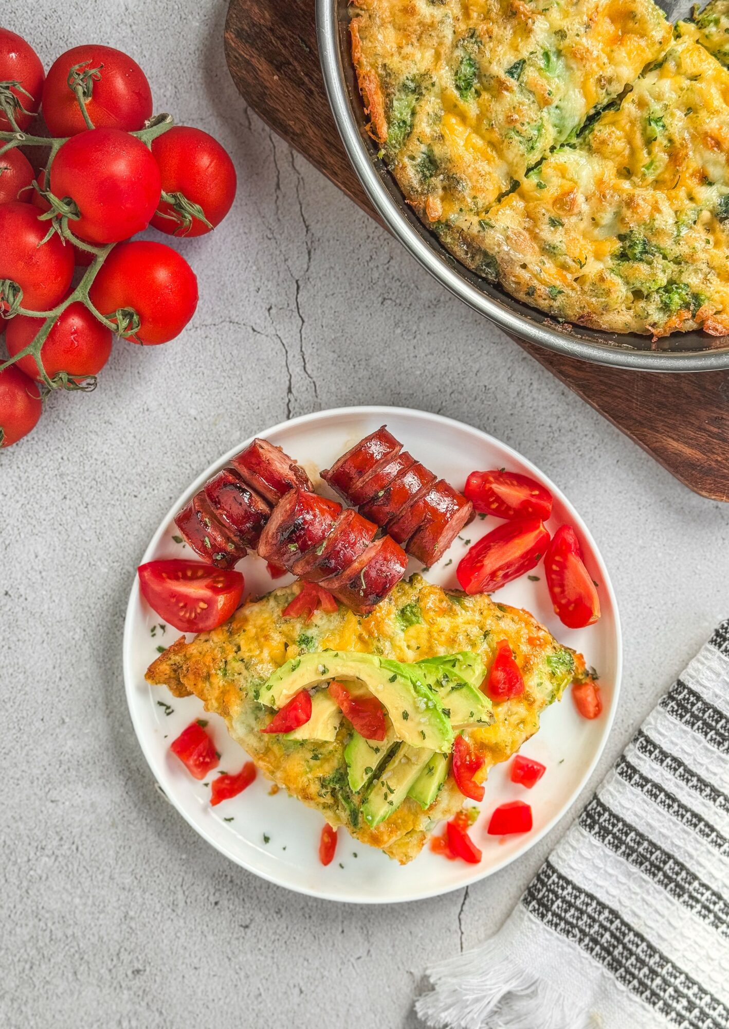 Plate with rolled meat, tomato slices, and a slice of vegetable frittata garnished with red bell pepper pieces, on a gray surface.