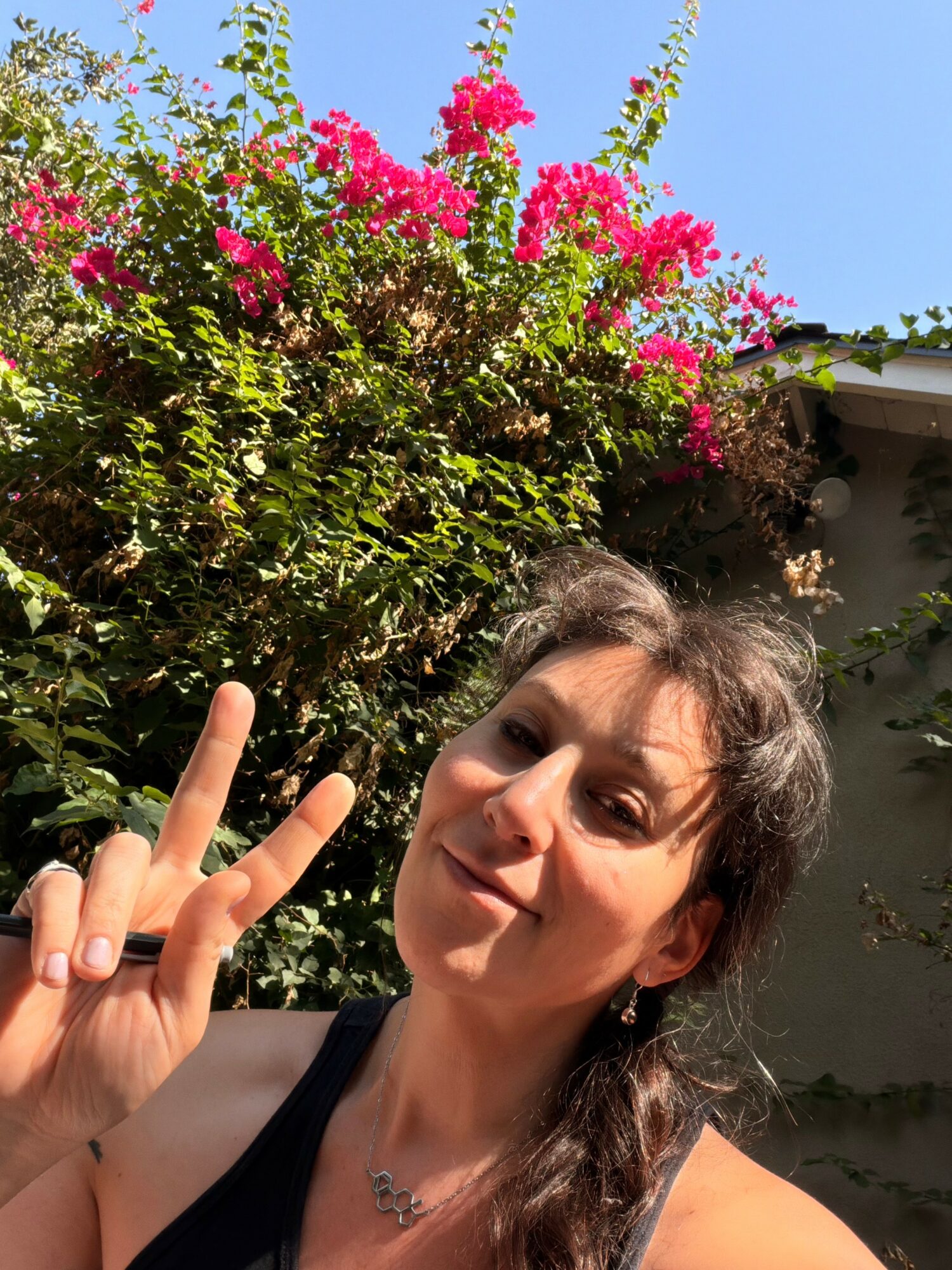 Woman with dark hair making a peace sign outdoors, pink flowering bush and blue sky in background.