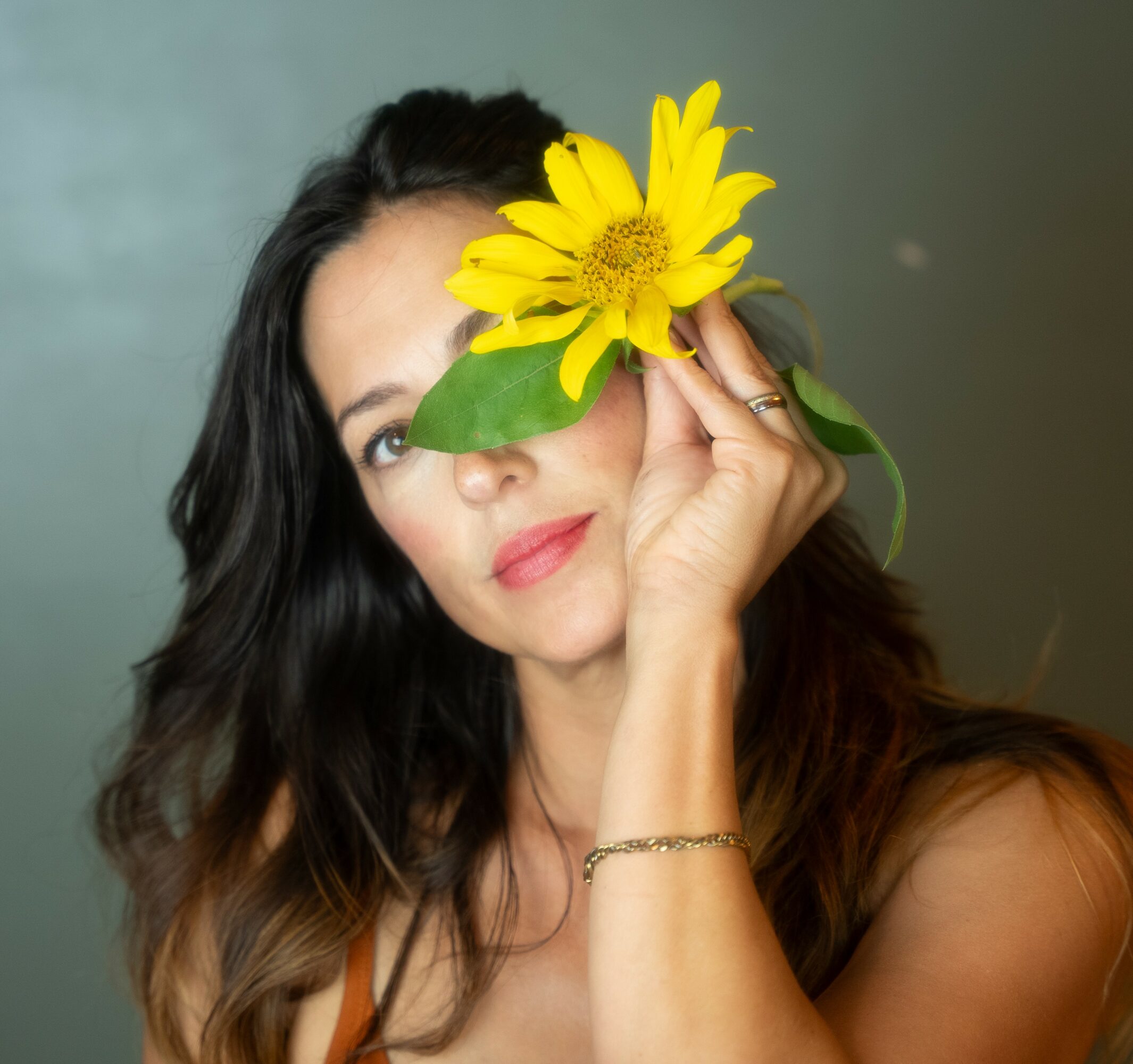 Woman holding a yellow flower in front of her eye, with dark wavy hair and a neutral background.
