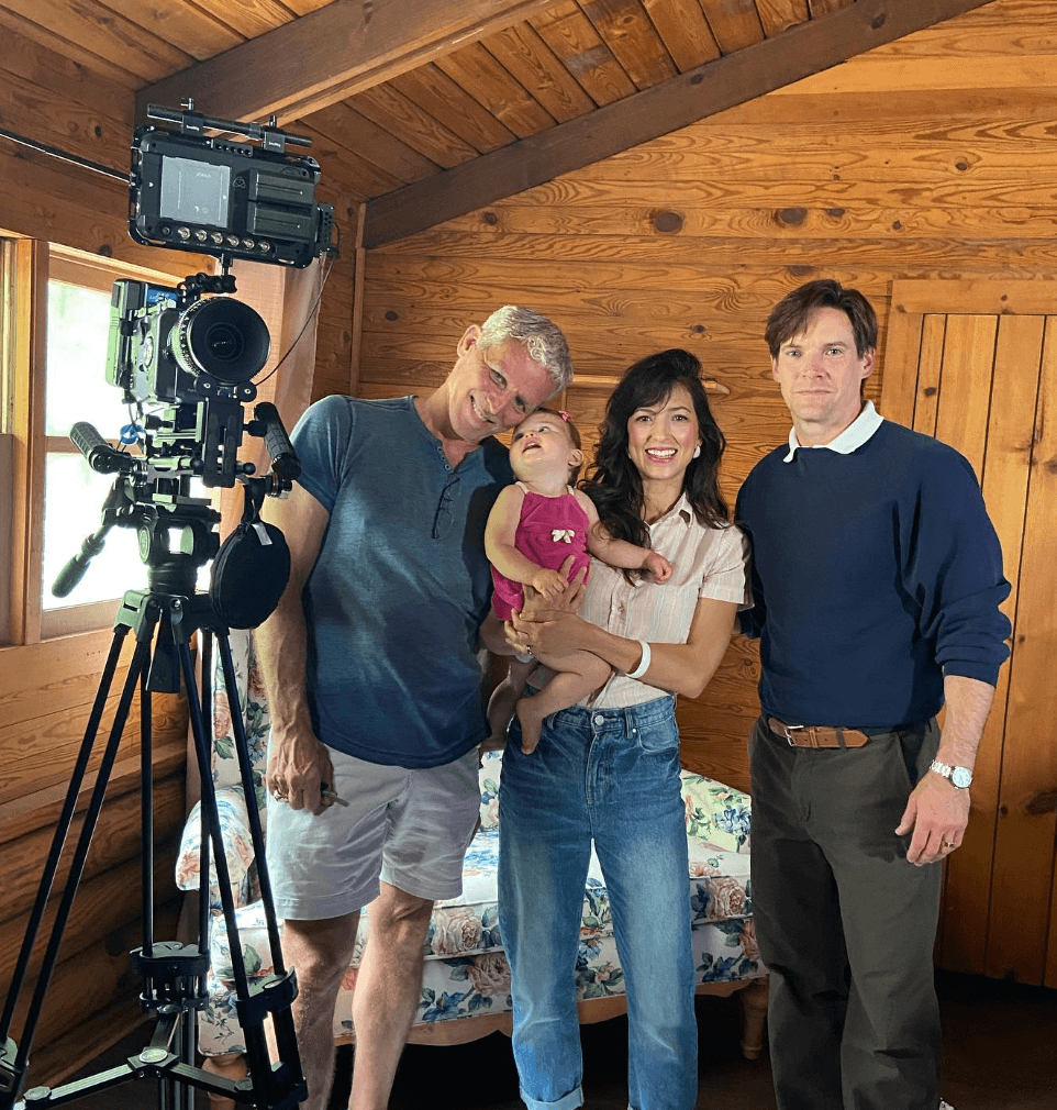 Three people and a doll in a wooden room, with a camera on a tripod. The woman holds the doll, smiling. The man and woman stand beside her. Word count: 20, confidence: 0.95