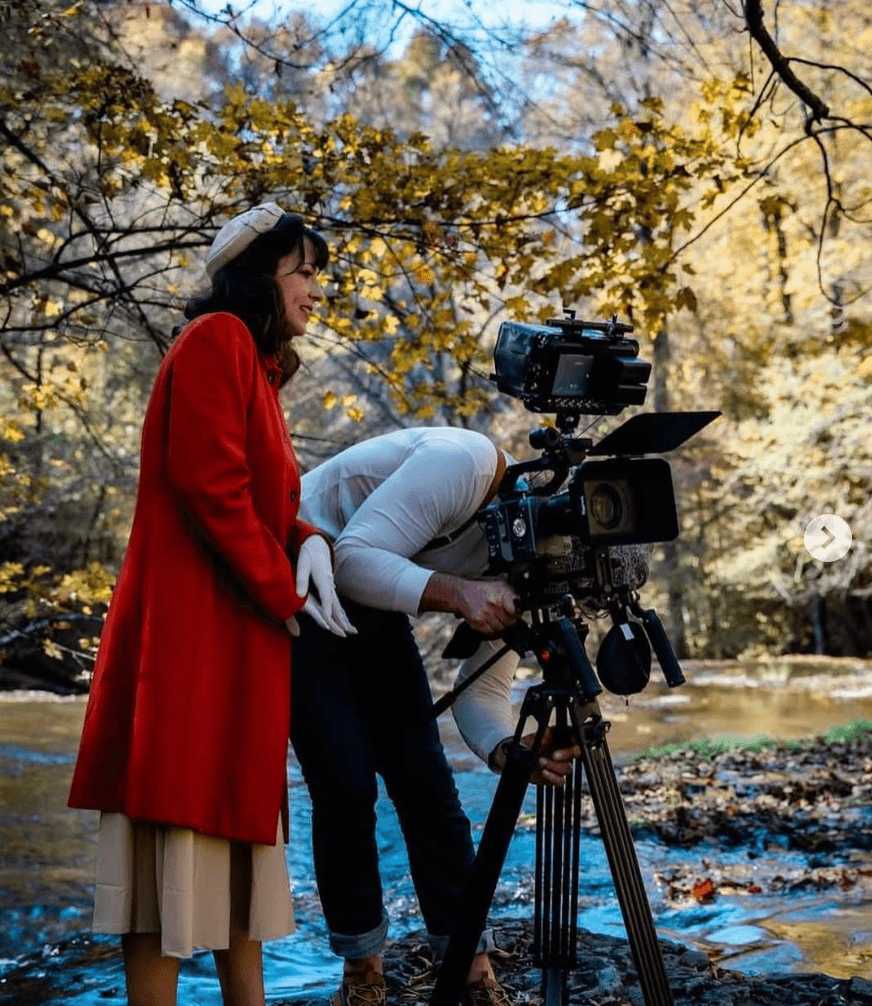 Two people filming outdoors in a forest with autumn leaves, one woman in a red coat and a person operating a camera.