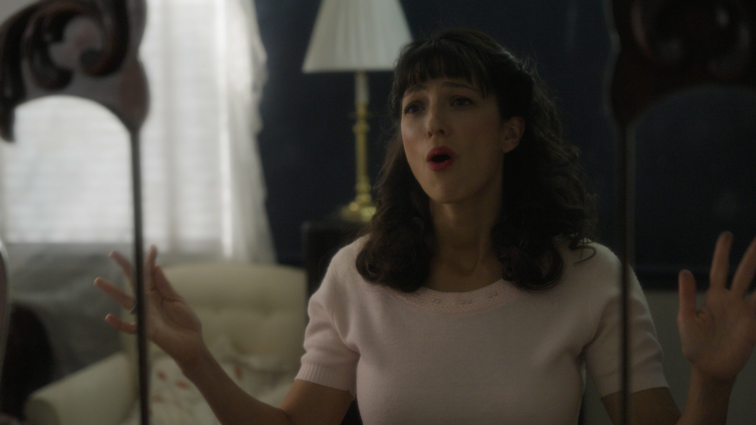 Young woman with dark hair and bangs gestures with hands in a room with a lamp and curtains.