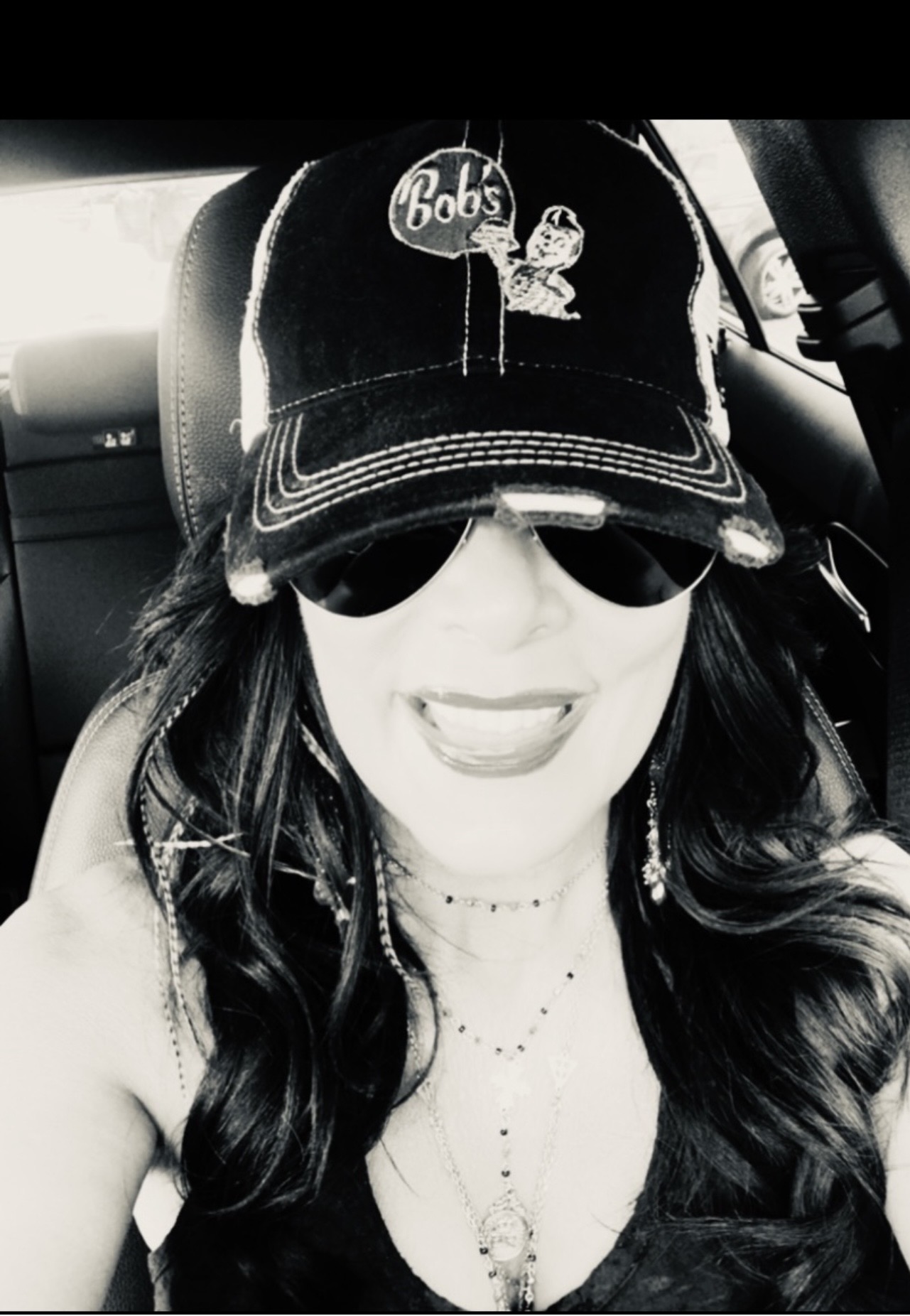 Woman wearing a baseball cap, sunglasses, and a necklace, smiling inside a vehicle.